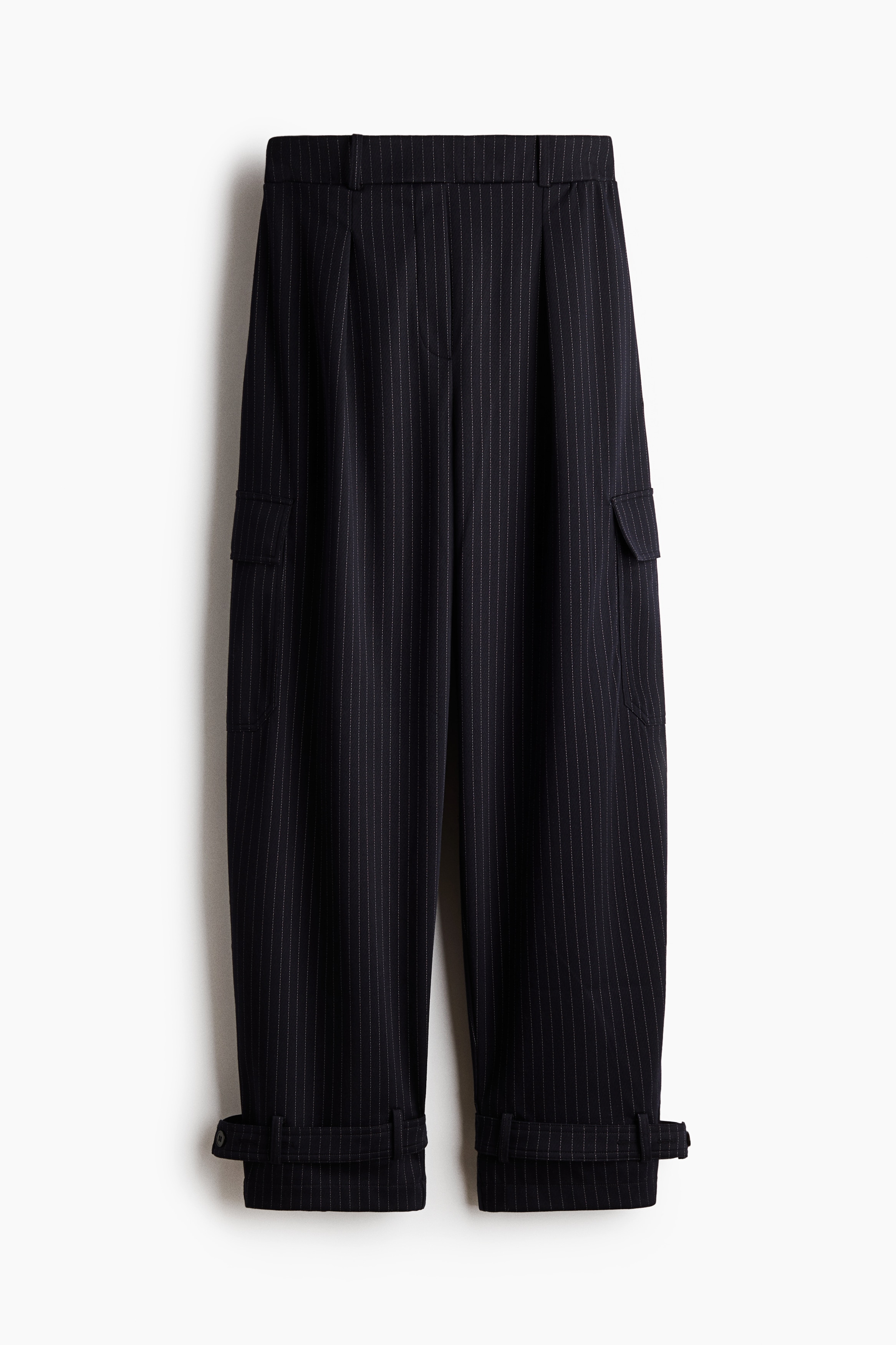 Loose-fit cargo trousers in dark pinstriped jacquard jersey are facing left, showcasing a wide-leg silhouette and a side flap pocket. The trousers sit at the waist and taper at the ankles with adjustable button-strap hems.
