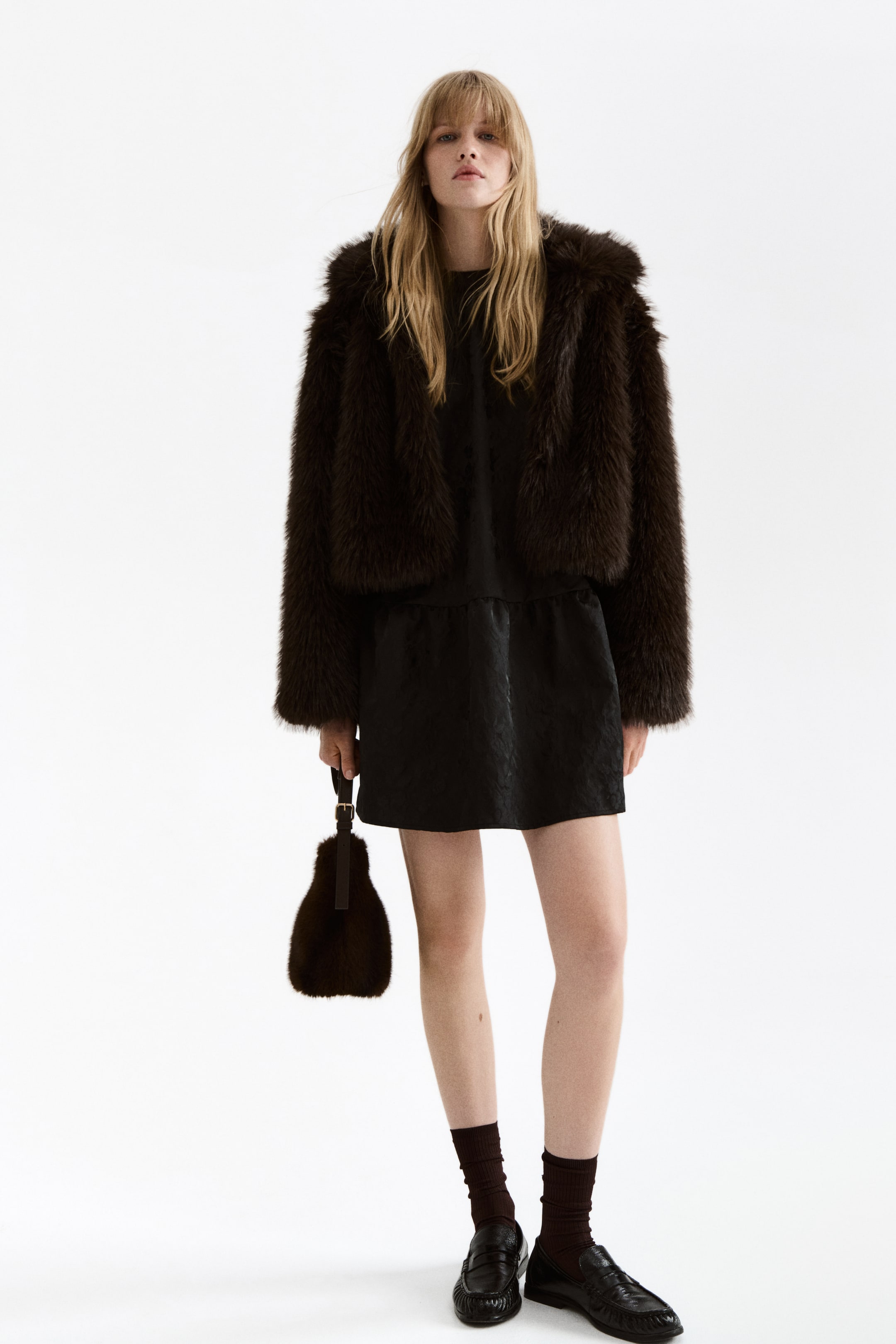 View larger image: A dark brown faux fur jacket is being presented front-facing, featuring a cropped length and a full, shaggy texture with long sleeves. It is layering loosely over a black short-sleeved dress.