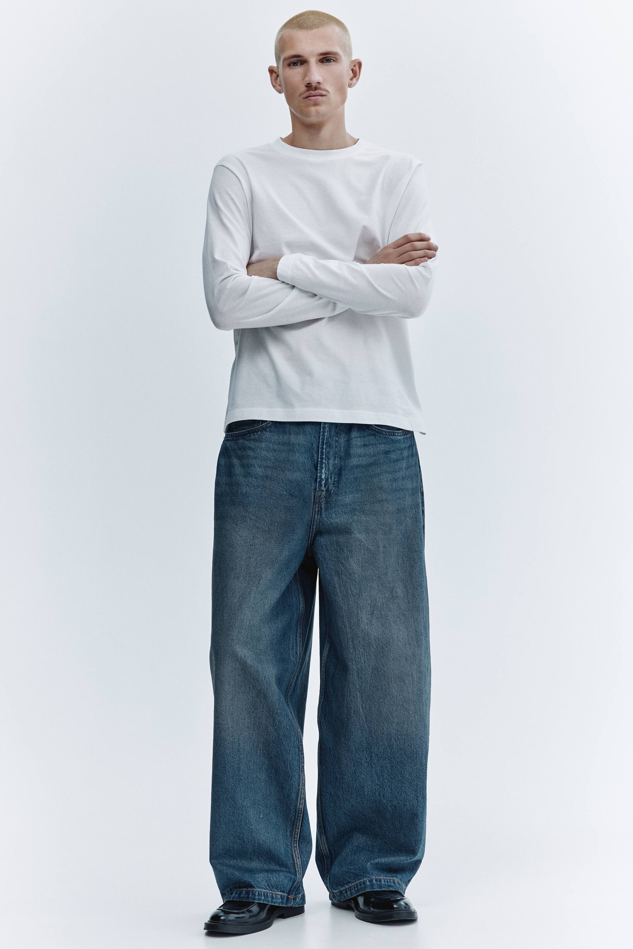 Преглед на по-голямо изображение: Представя се бяла тениска с дълъг ръкав и кръгло деколте, с гладка текстура и фина ребриста лента по деколтето. Тя е комбинирана със сини дънки с широк крачол, с умерено измиване, по‑светли избледнявания и леко струпване върху обувките.