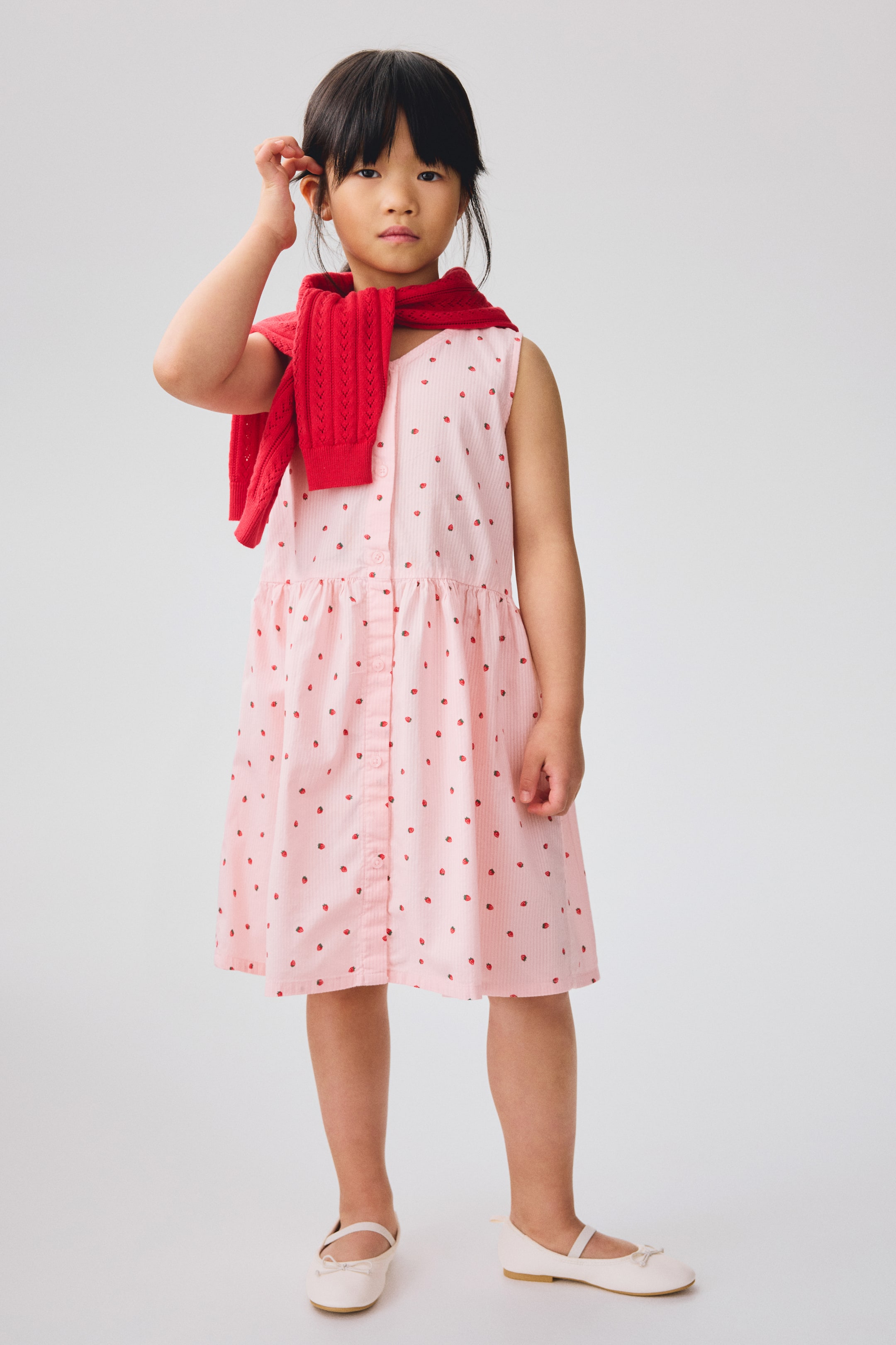 View larger image: The pink sleeveless dress showcases white vertical stripes and a red strawberry print. It is crafted with a button-front placket and a gathered waist, forming a full skirt that falls above the knees.
