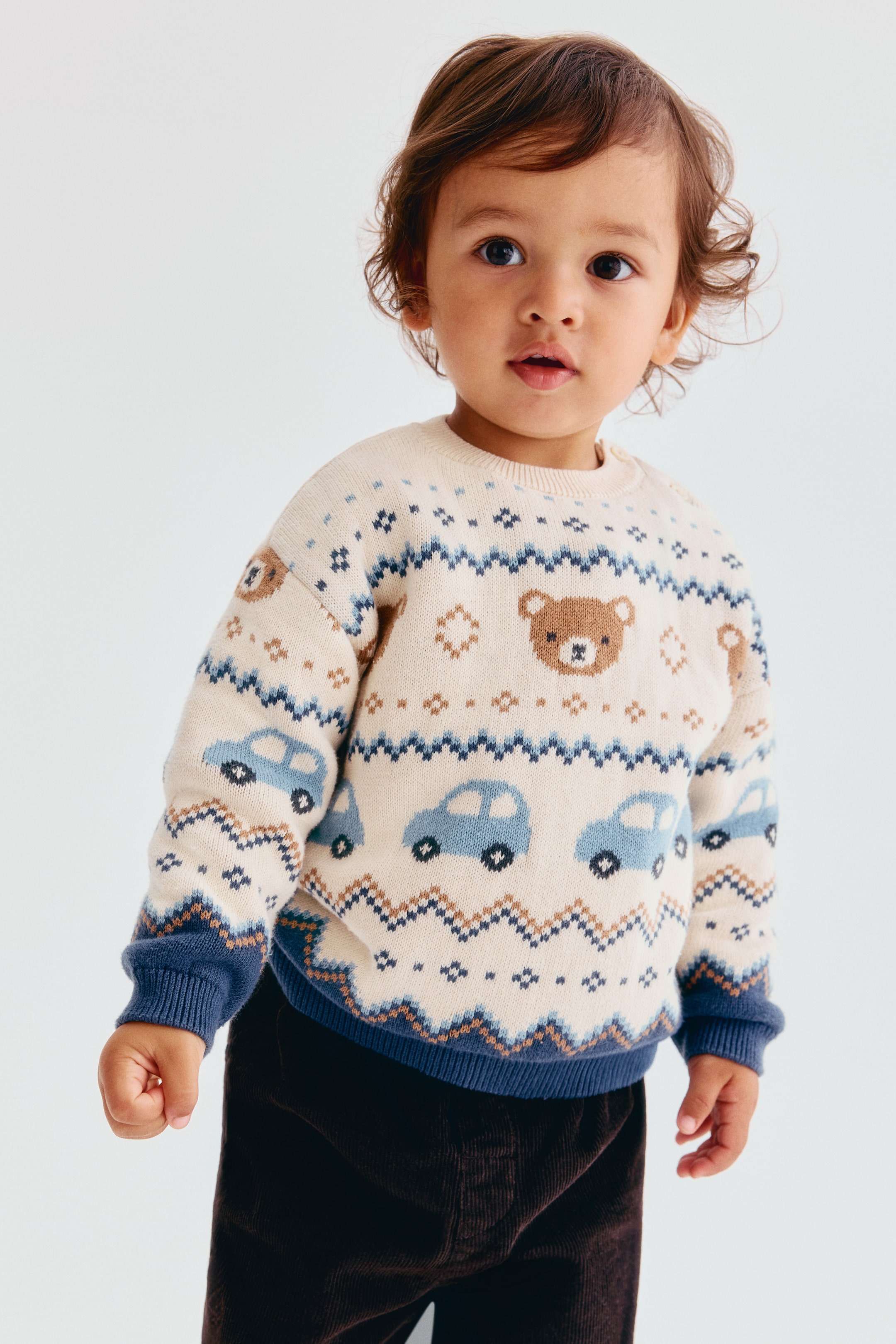 Grösseres Bild ansehen: Ein cremefarbener Jacquard-Strickpullover aus Baumwolle wird von vorne gezeigt und weist horizontale Muster mit blauen Autos, braunen Teddybären und geometrischen Zickzacklinien auf. Der Pullover präsentiert sich in einer lockeren Passform mit überschnittenen Schultern, einem gerippten cremefarbenen Ausschnitt sowie kontrastierenden blauen Rippbündchen an Ärmeln und Saum.