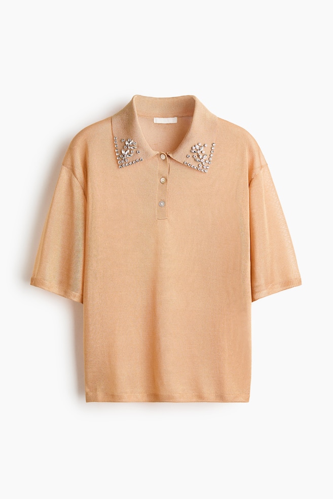 A peach-colored knit polo shirt is shown facing front, featuring a spread collar adorned with floral-patterned rhinestone embellishments. The shirt is designed with a three-button placket, dropped shoulders, and relaxed short sleeves in a fine-knit texture. - 1