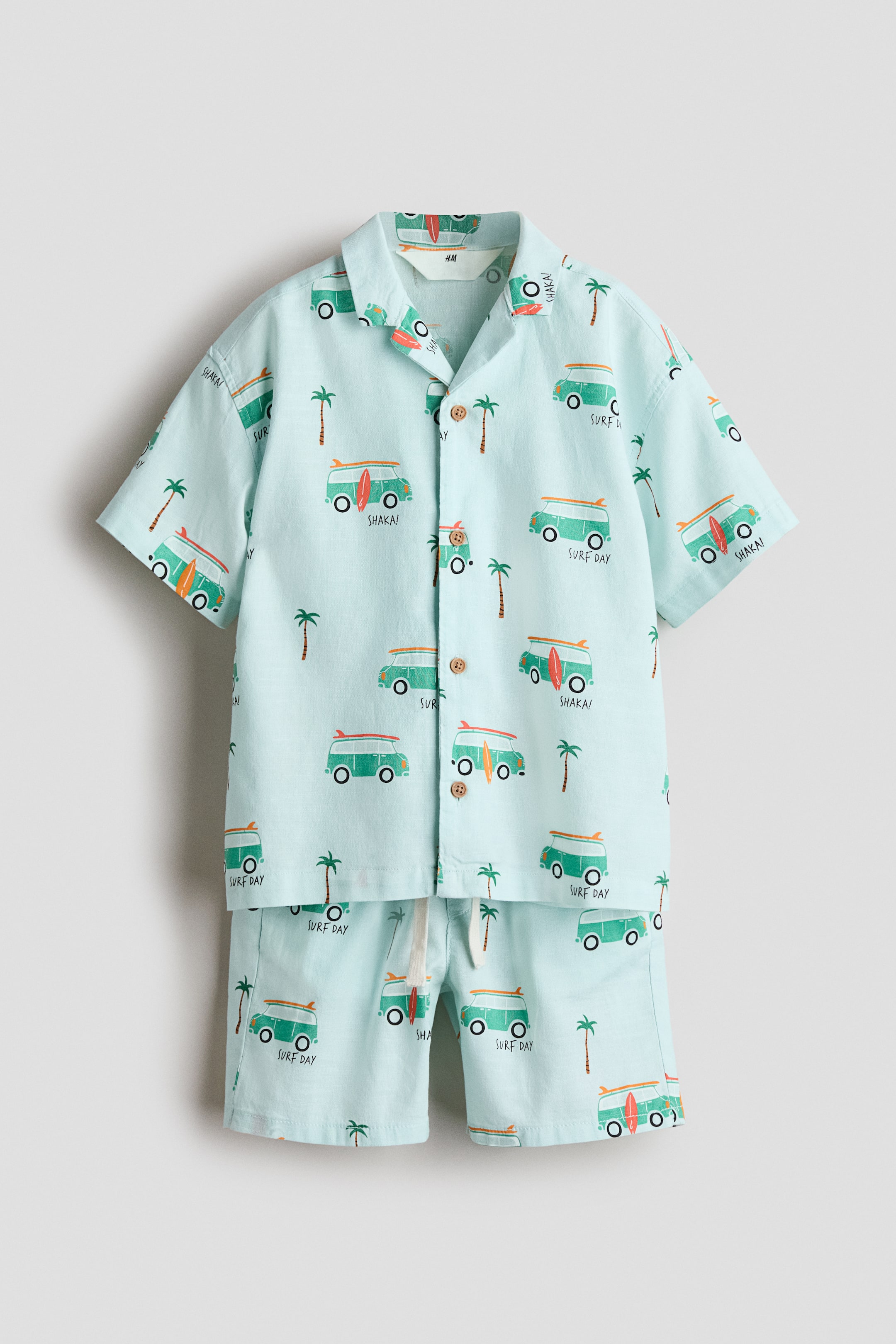 View larger image: A light blue two-piece set, comprising a short-sleeved shirt and matching shorts, showcases a repeating pattern of green surf vans, palm trees, and the words "SURF DAY" and "SHAKA!". The shirt features a camp collar and a front button fastening with light brown buttons, while the shorts boast a white drawstring waist.