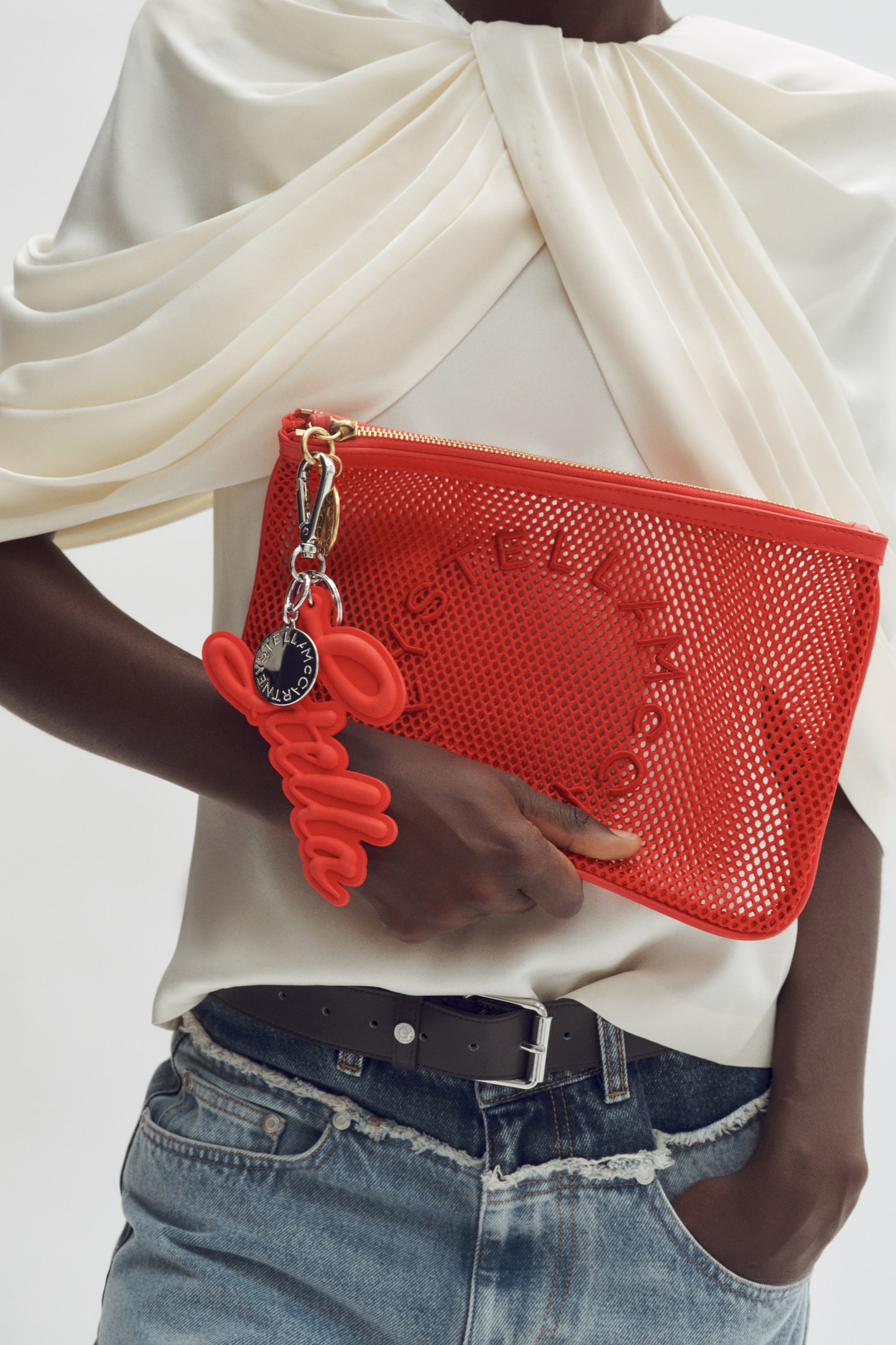 View larger image: A red mesh clutch bag is being held, angled slightly to the right, featuring a solid border, a gold zip closure, and Stella McCartney lettering visible on its front. A matching red sculptural keychain, featuring raised Stella McCartney lettering and a circular metallic tag, is hanging from the zip pull.