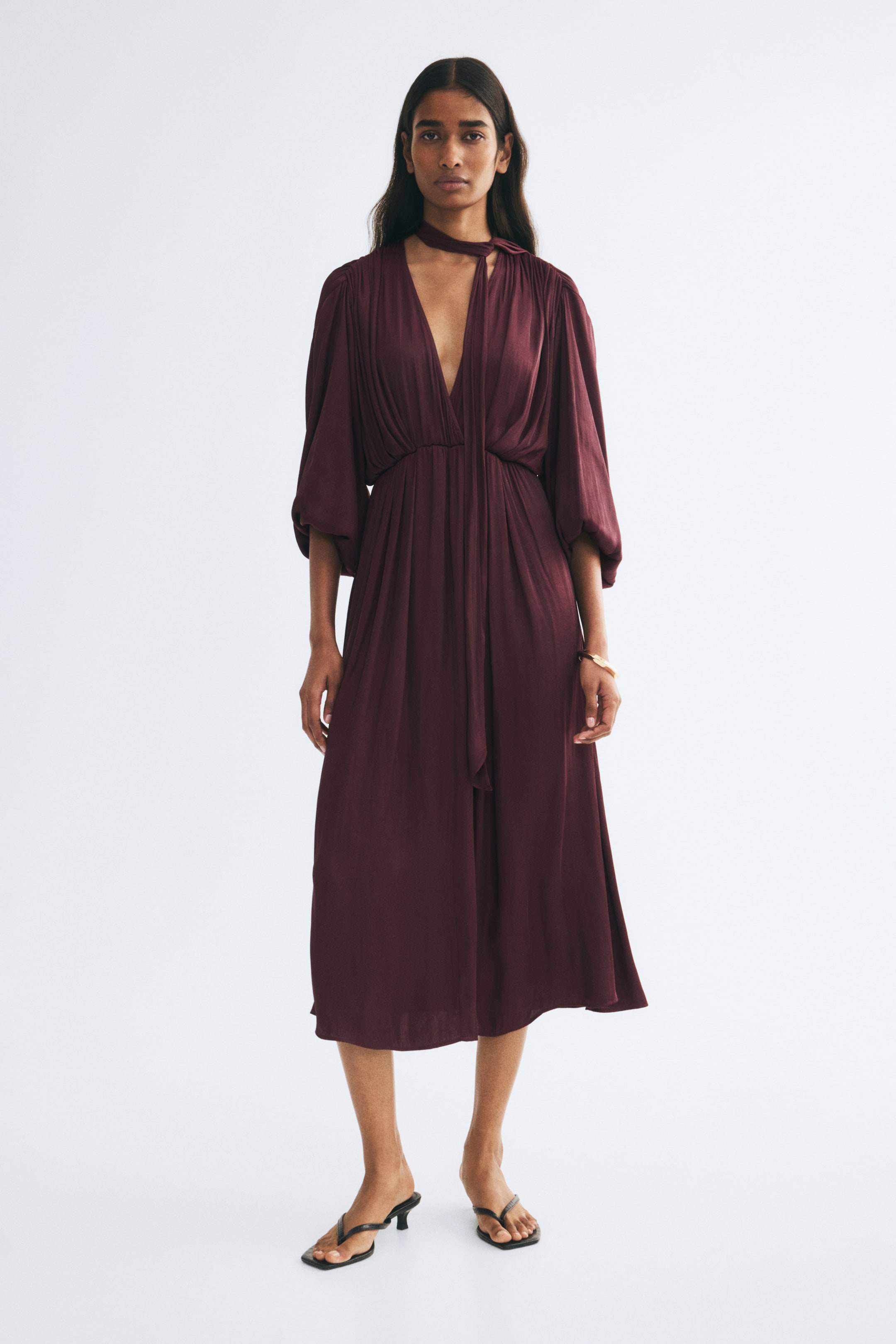 View larger image: The model is wearing a burgundy midi dress in draping satin twill, featuring a deep V-neckline, a high scarf collar, and long, gathered balloon sleeves. The dress is styled with an elasticated waist and a gently flared skirt, and is paired with black thong sandals.