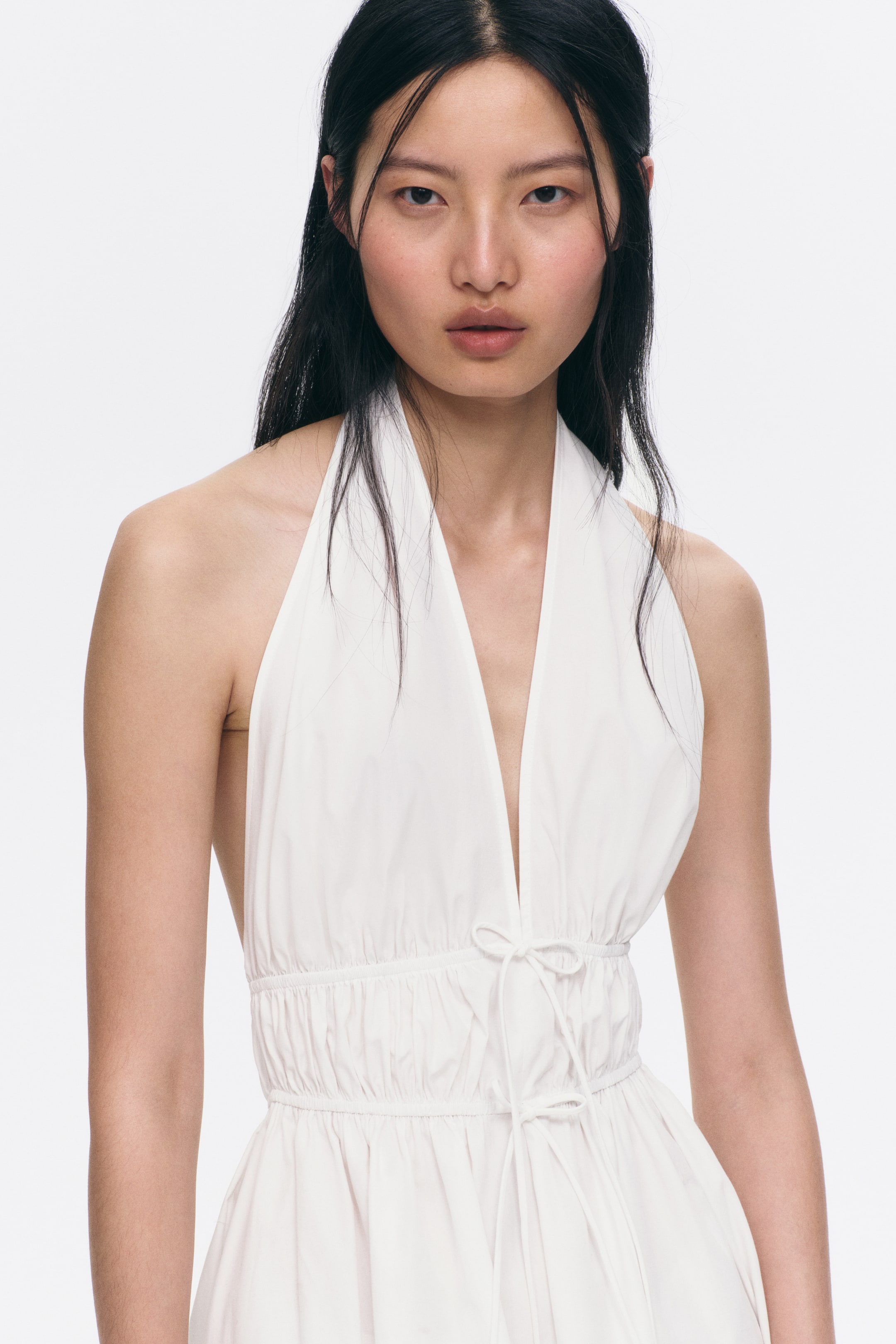 View larger image: A white halter-neck dress featuring a deep V-neckline and a gathered waist, with a tie detail visible at the front. The skirt flows outwards from the waist gathers.