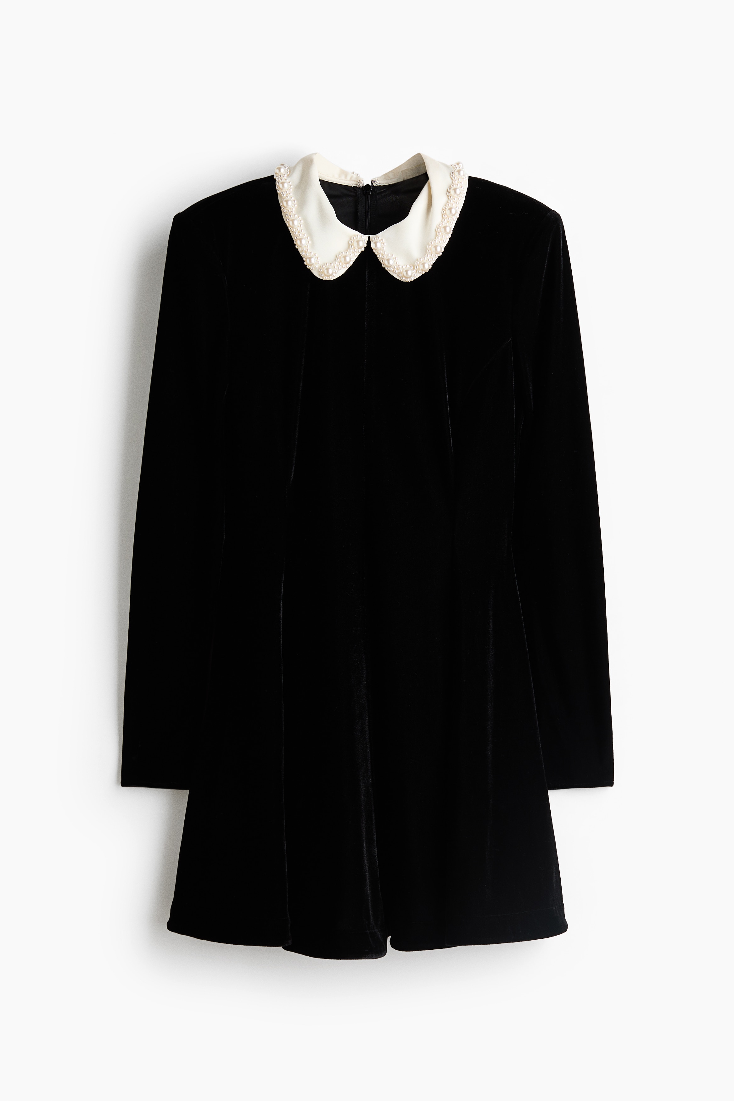 A short, dark velour dress with long sleeves, a fitted bodice and a contrasting white beaded collar is worn by the model. Standing with hands on hips to emphasise the flared skirt, the model pairs the dress with black heeled sandals.