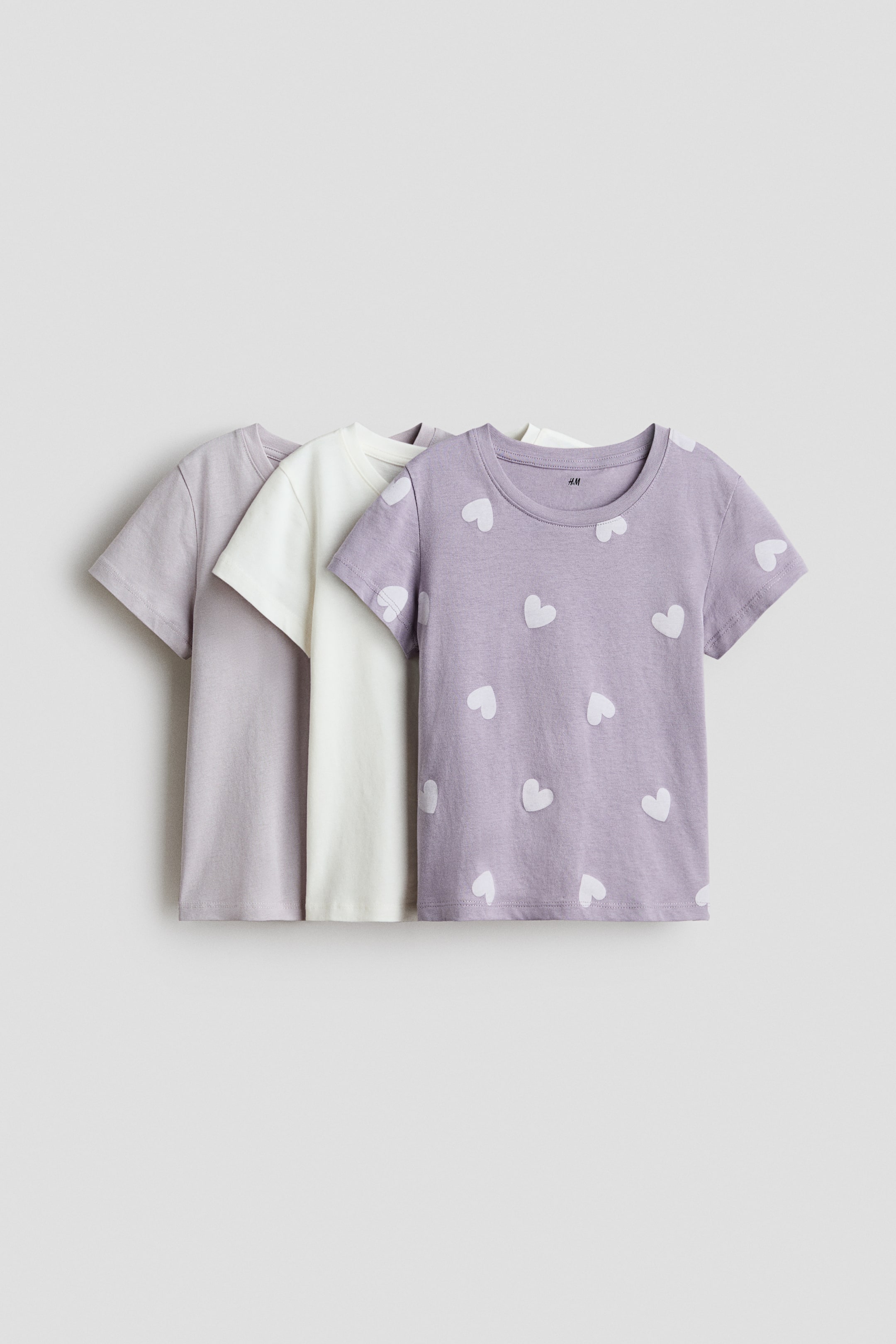 View larger image: Three t-shirts are positioned facing forward, slightly layered. The front t-shirt is a light purple adorned with white heart patterns, featuring a round neckline and short sleeves. An off-white t-shirt is layered behind it, and a solid light lavender t-shirt is layered further back, both also showcasing round necklines and short sleeves.