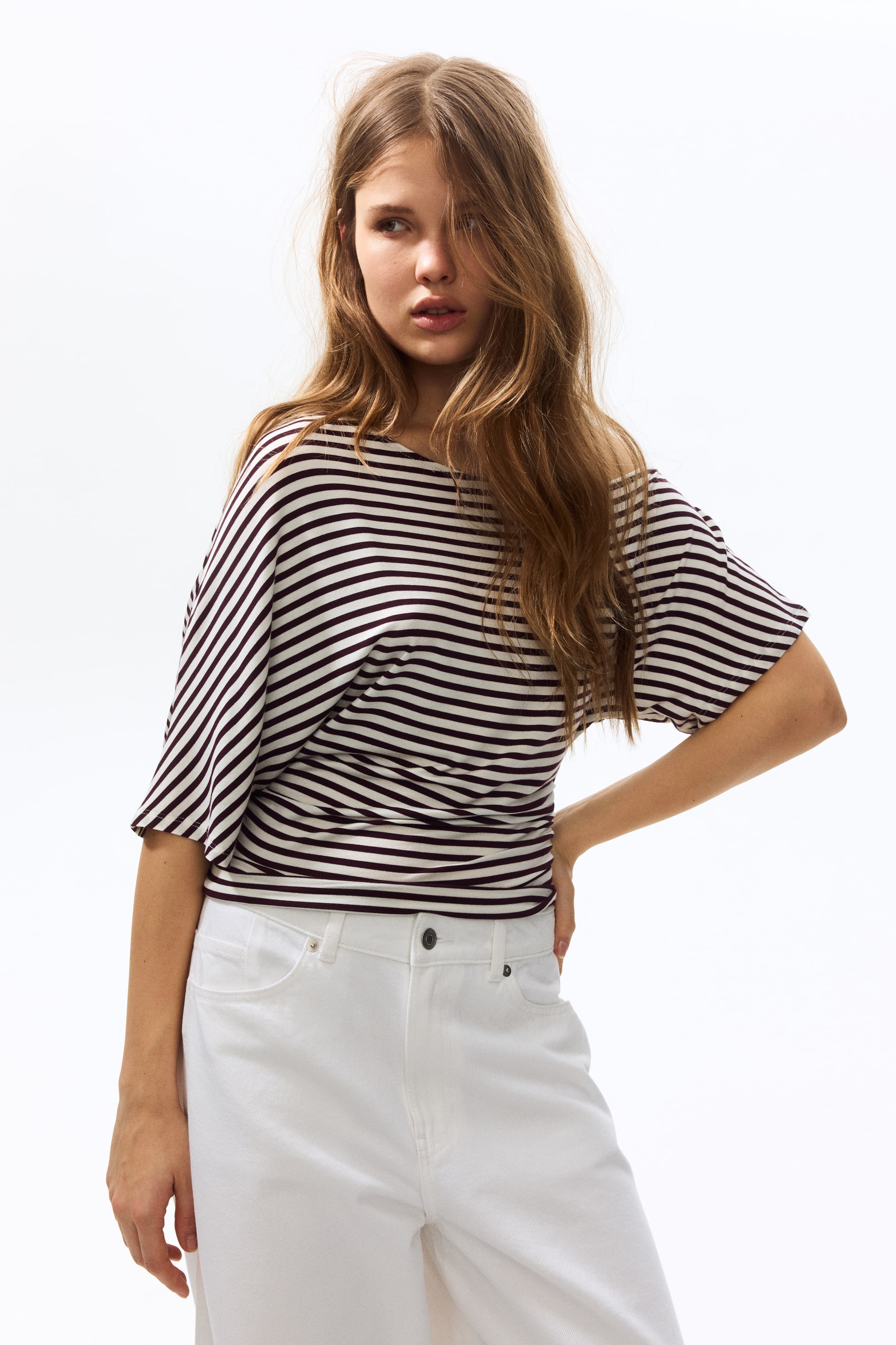 Ver imagem maior: Um top às riscas apresenta riscas horizontais em castanho escuro e branco sujo, um decote barco largo e mangas dolman largas, com a bainha franzida a assentar na cintura.