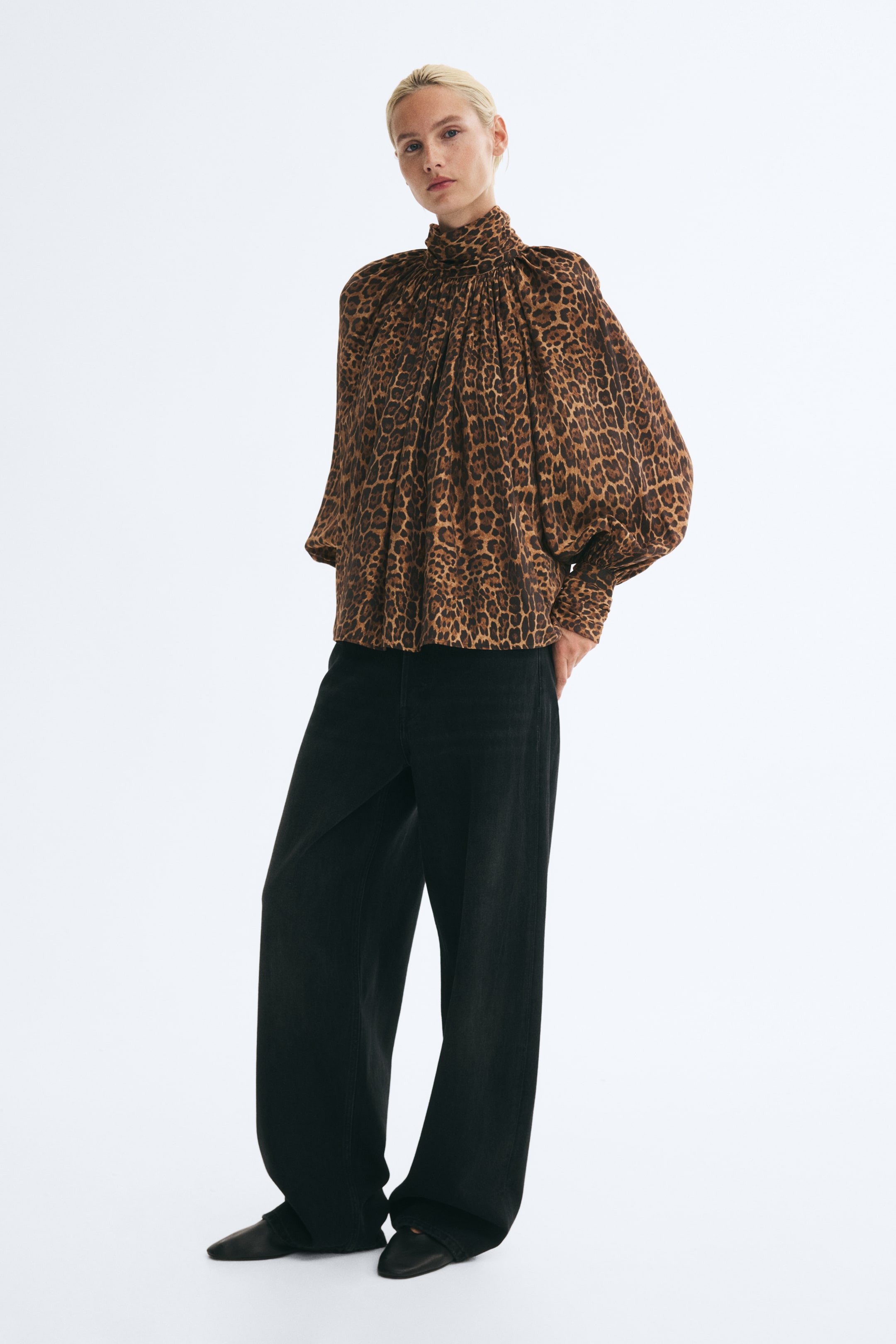 Zobraziť väčší obrázok: Modelka má oblečenú oversized blúzku s leopardím vzorom, s vysokým nariaseným stojatým golierom so skladmi a objemnými balónovými rukávmi raglánového strihu so širokými manžetami. Blúzka je skombinovaná s čiernymi nohavicami so širokými nohavicami.
