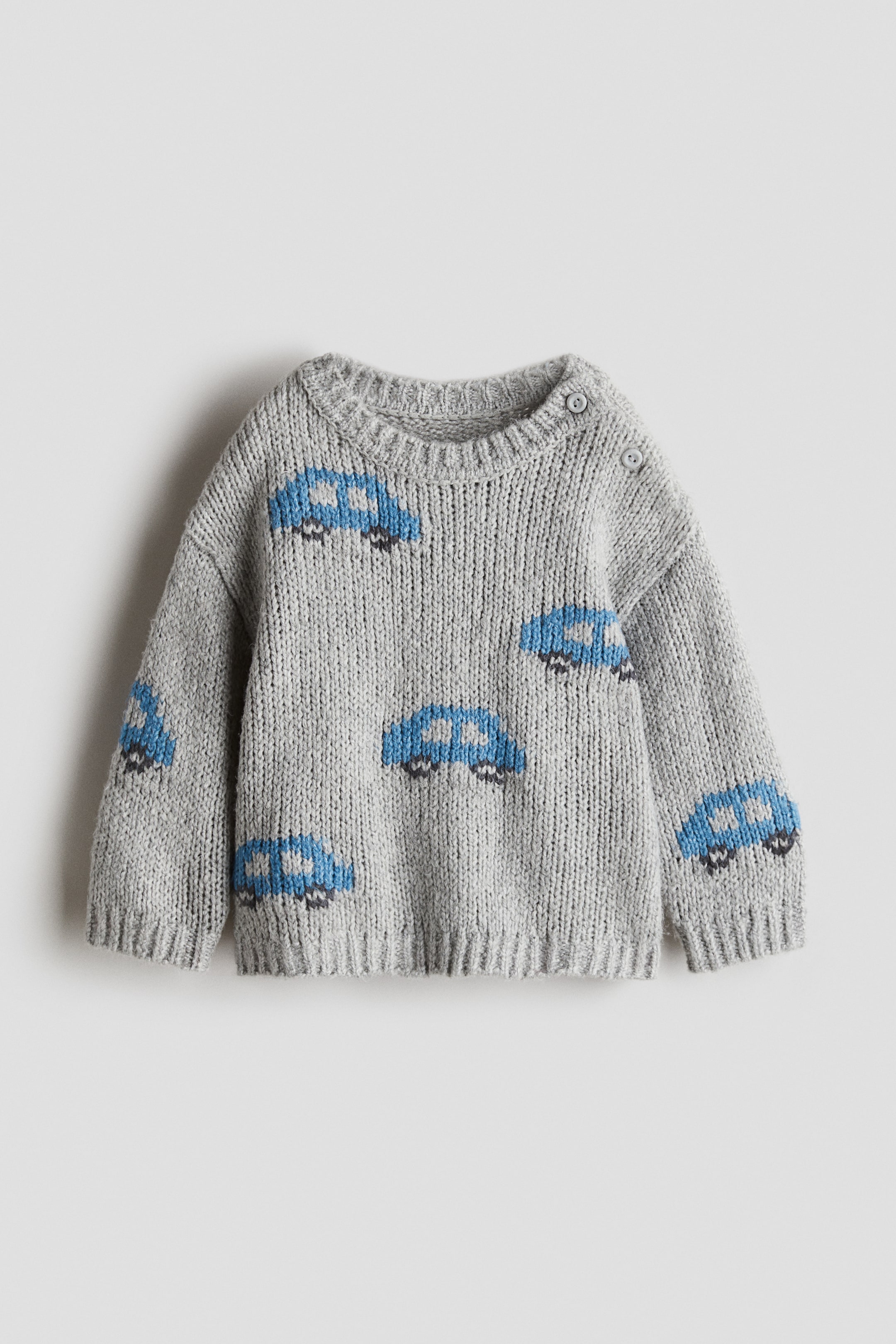 Se større billede: En grå strikket sweater er mønstret med blå biler og sorte hjul, og har en ribbet rund hals, ribbede manchetter og en ribbet kant. Den lukkes med to små grå knapper på venstre skulder.