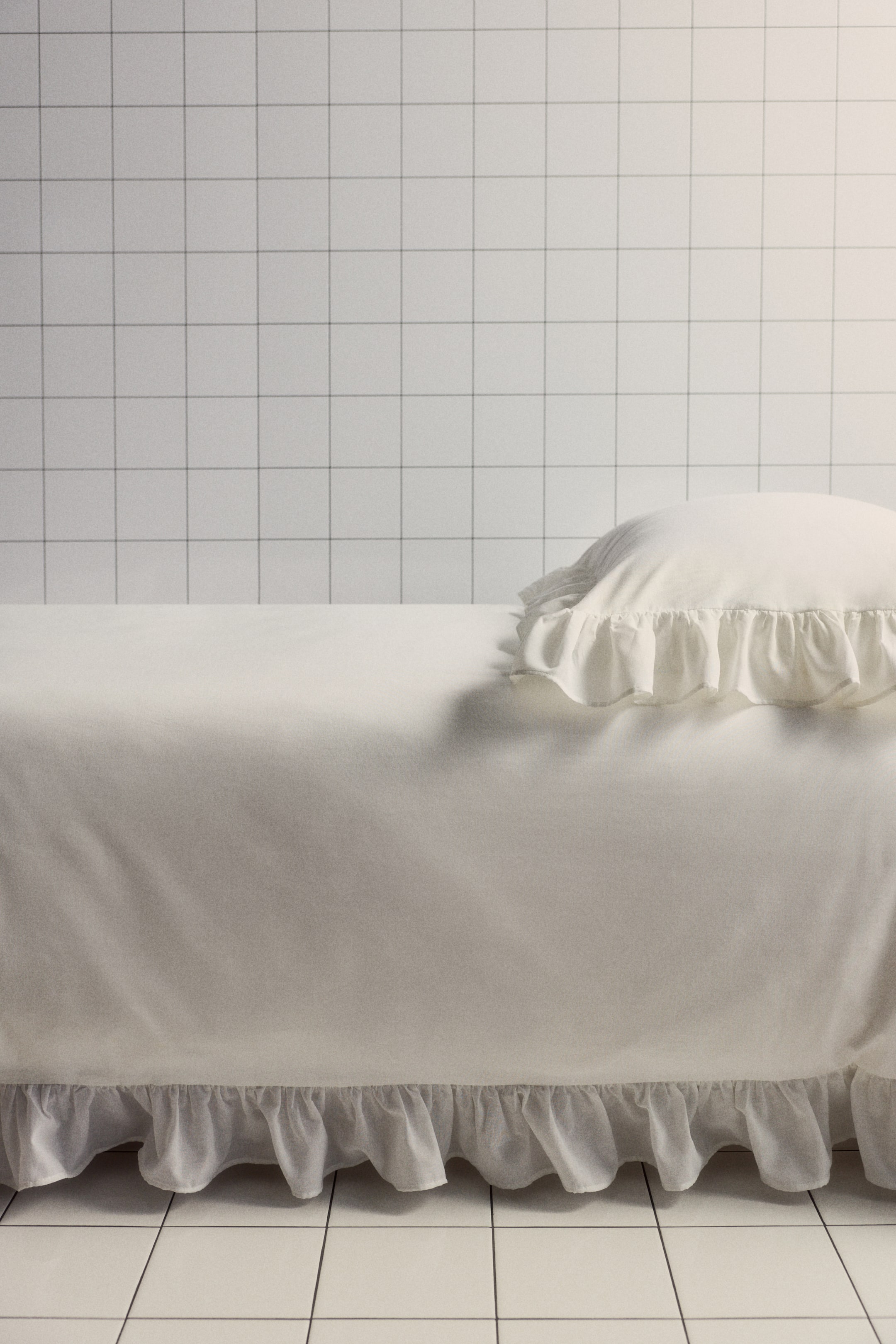 拡大画像を表示: オフホワイトのベッドスカートが側面を見せており、ベッドの側面を覆う滑らかなパネルと、ギャザーを寄せたフリルの裾が特徴です。マッチするオフホワイトのフリル縁の枕がベッドに置かれています。