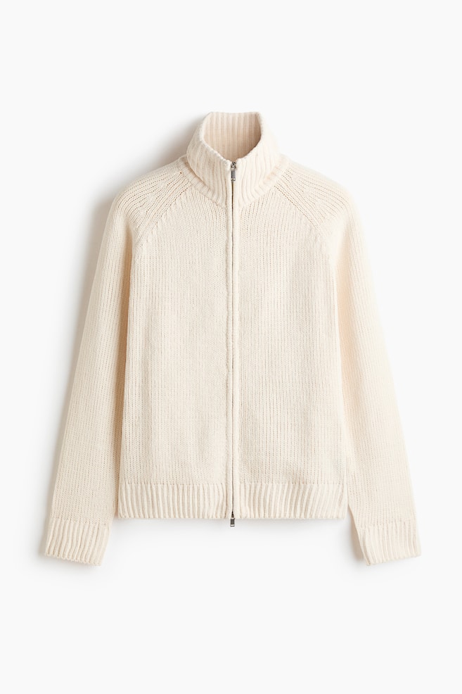 Un chandail zippé en tricot de couleur crème est présenté de face, avec une fermeture éclair pleine longueur et un col montant côtelé. Il est également doté de poignets côtelés, d'un ourlet côtelé et de manches raglan. - 1