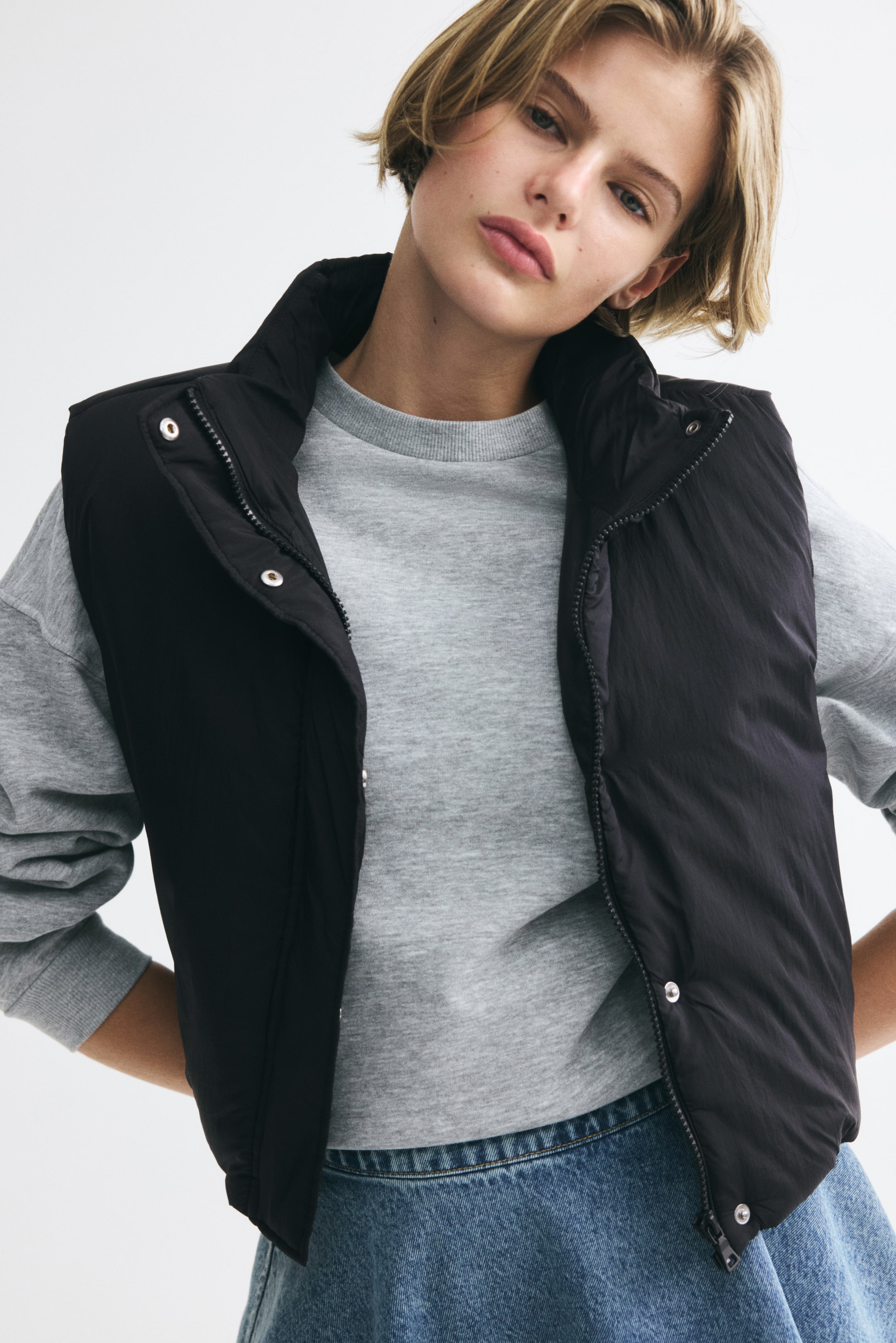 Voir une image plus grande: Un gilet matelassé noir en nylon, doté d’un col montant et d’une ouverture à l’avant avec des boutons-pression argentés, est porté par le mannequin. Le gilet est superposé à un sweatshirt gris à manches longues et agencé avec un bas en denim bleu.