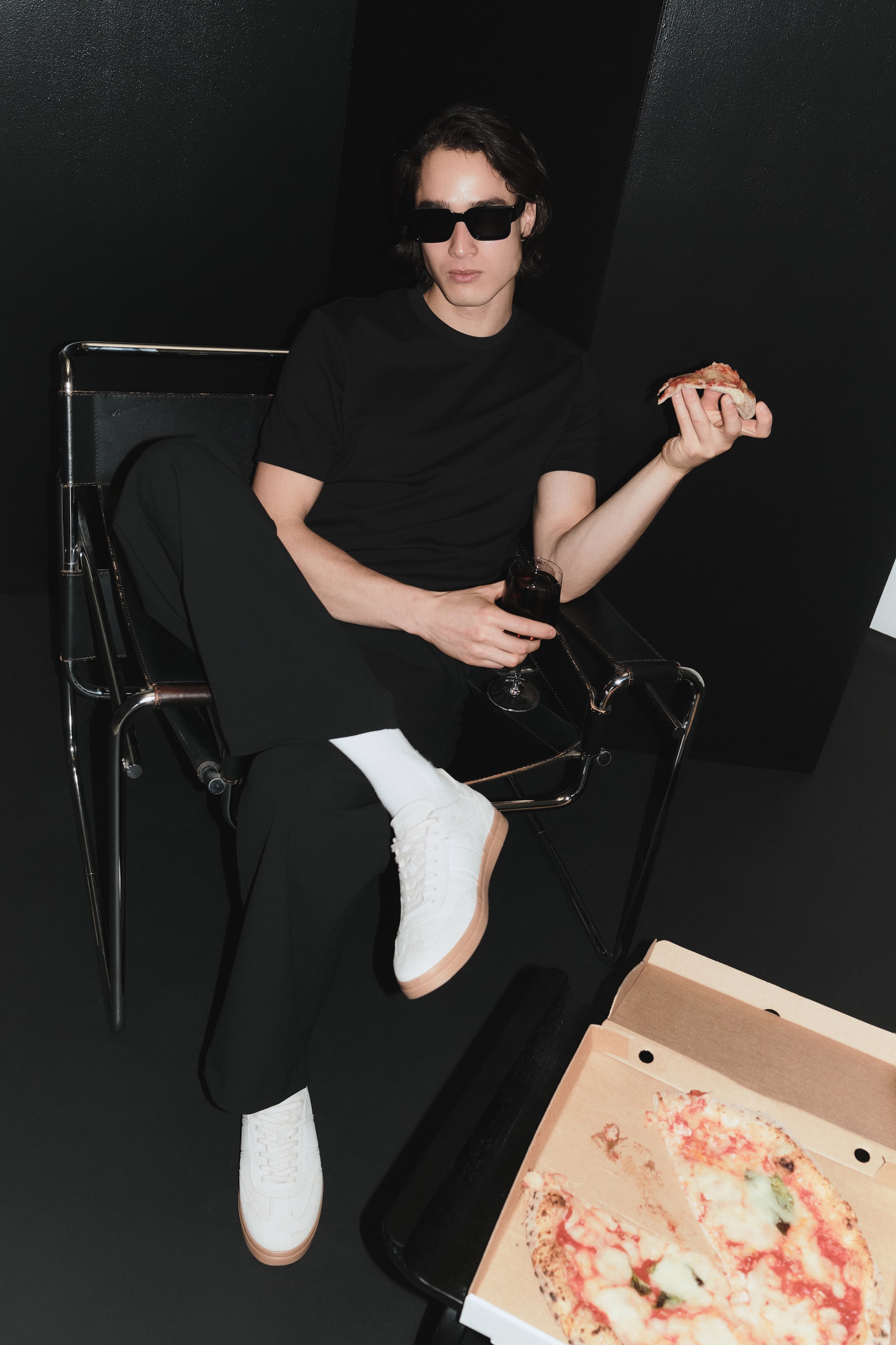 Größeres Bild ansehen: Ein schlichtes schwarzes T-Shirt mit Rundhalsausschnitt ist mit einer locker sitzenden schwarzen Hose und weißen Low-Top-Sneakern kombiniert, die hellbraune Sohlen und weiße Schnürsenkel aufweisen. Zwischen Hose und Sneakern sind weiße Socken sichtbar, während eine schwarze, rechteckig gerahmte Sonnenbrille auf dem Gesicht sitzt.