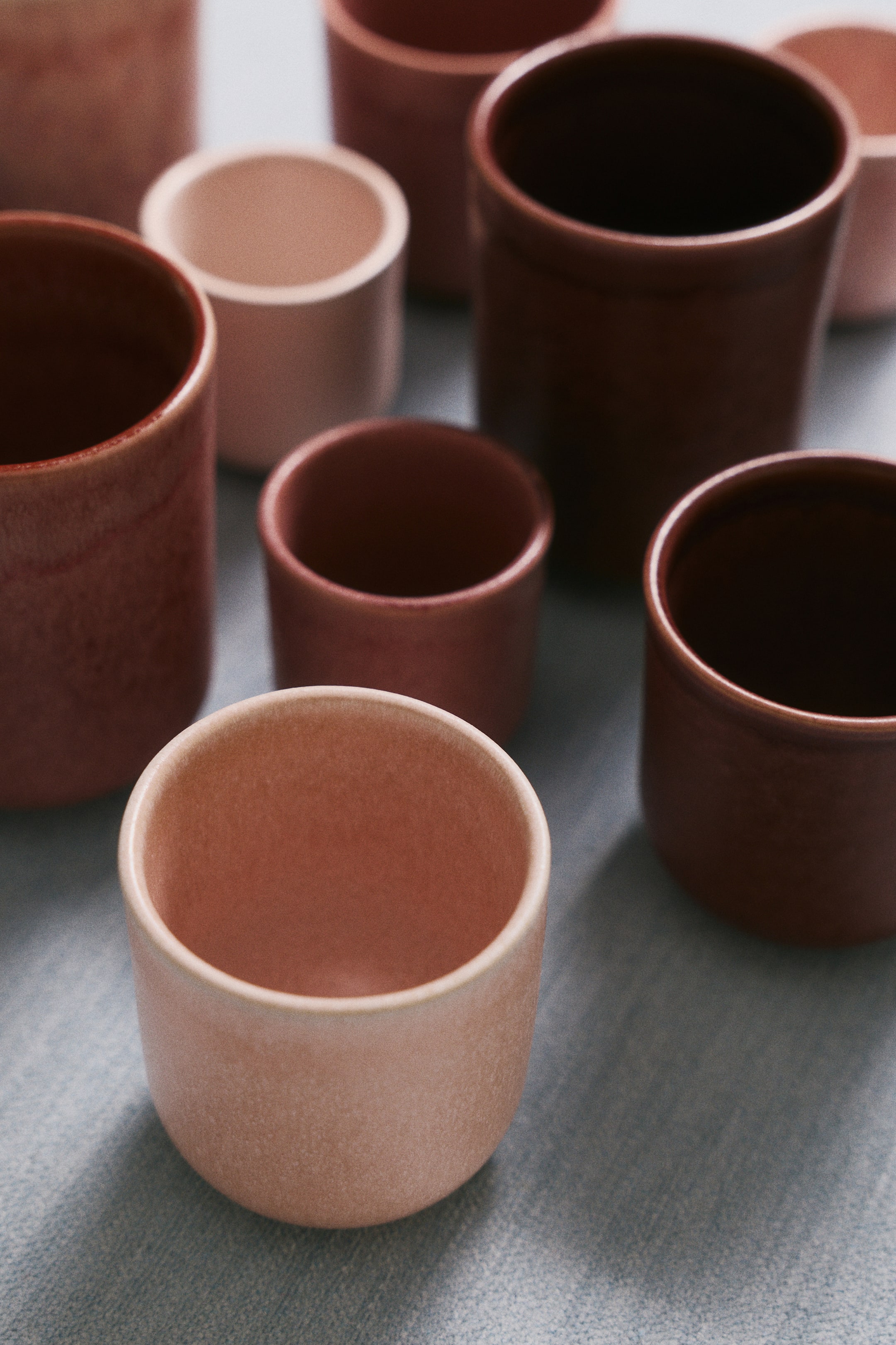 View larger image: A collection of handleless stoneware mugs in varying sizes and earthy shades of dusty pink, terracotta, and dark brown showcases a rustic, cylindrical design. The high-angle view highlights the unique, variegated reactive glaze and smooth, rounded rims of the vessels.