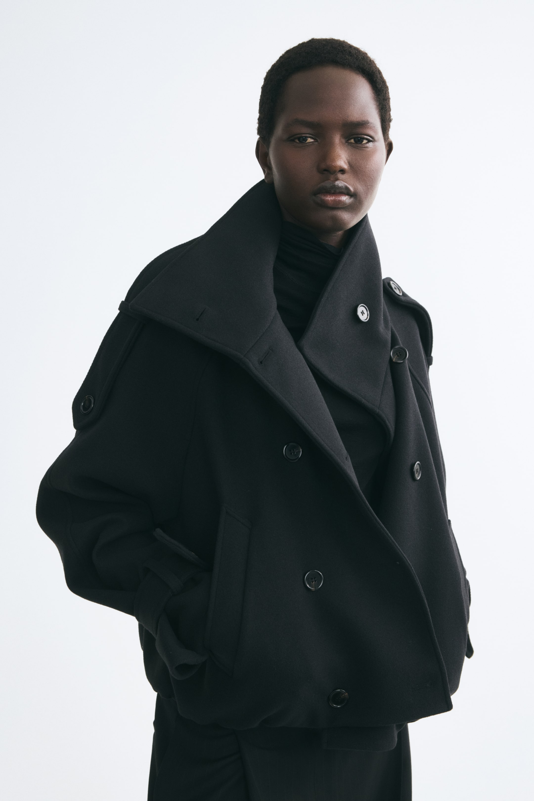 Ver imagem maior: Um casaco preto cruzado em tecido feltro é usado pela modelo sobre uma camisola de gola alta preta. O casaco oversized apresenta uma gola larga, dragonas abotoadas e bolsos embutidos à frente, com a modelo a colocar uma mão no bolso.