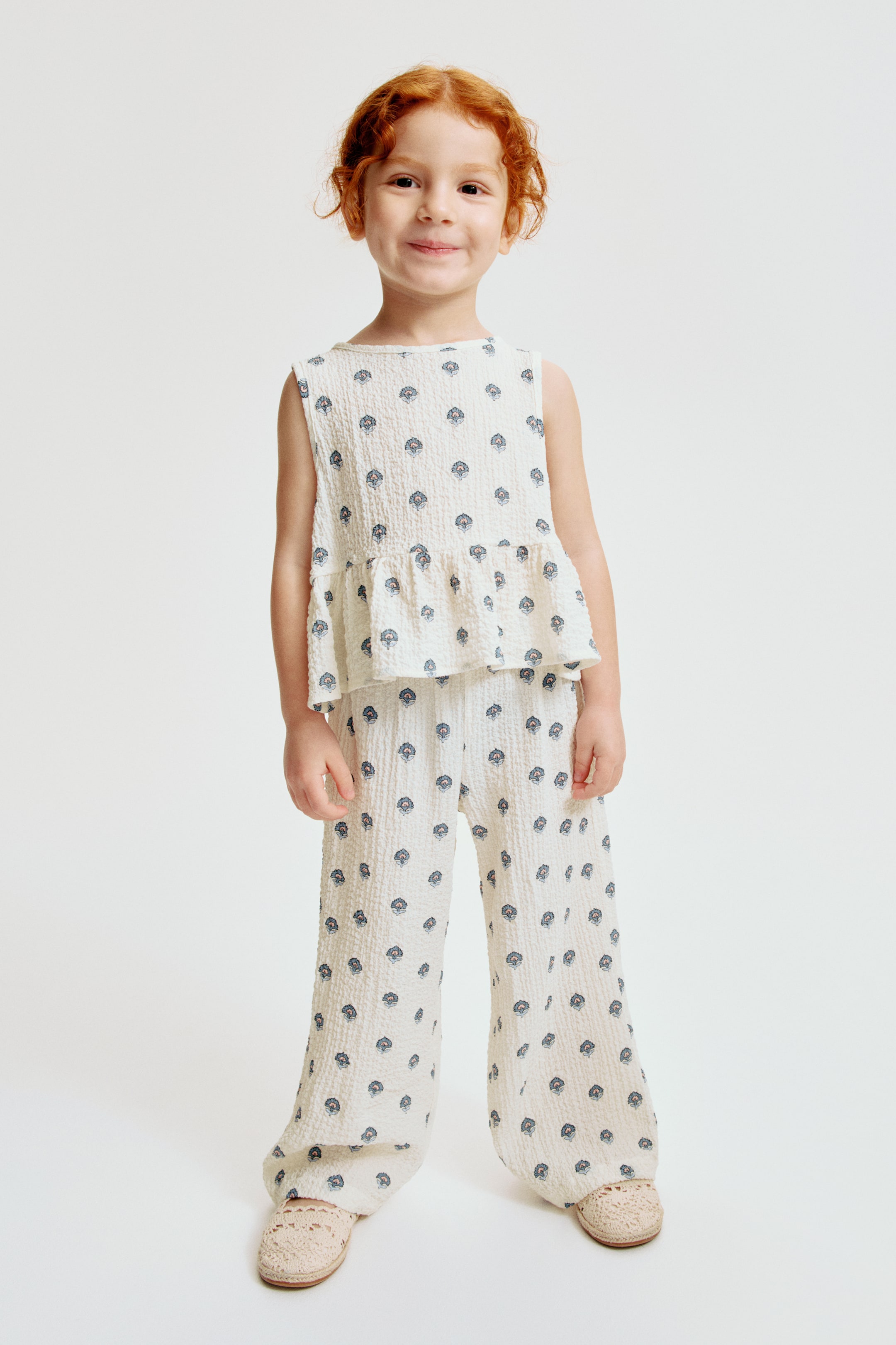 View larger image: A two-piece kids' set featuring an off-white textured fabric adorned with a repeating blue and red floral pattern. The sleeveless top is crafted with a round neckline and a peplum hem, while the matching full-length pants showcase a wide-leg style.