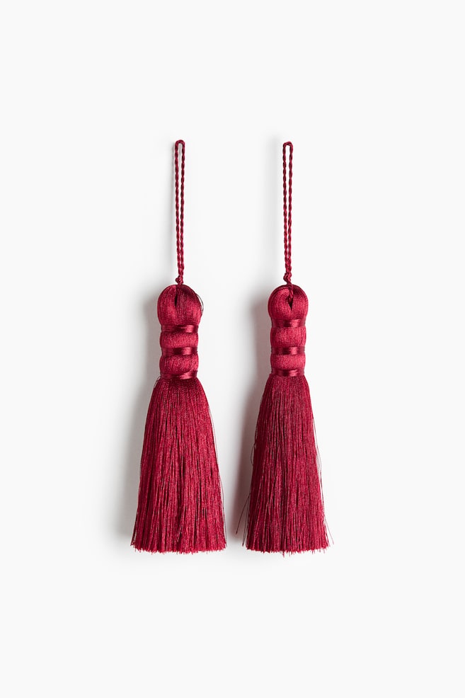 Une paire de pompons rouge foncÃ© est suspendue cÃ´te Ã  cÃ´te, orientÃ©e vers lâavant. Chaque pompon prÃ©sente en haut une fine boucle de cordon torsadÃ© suspendue, une tÃªte bulbeuse segmentÃ©e enveloppÃ©e de bandes horizontales de fil assorti, ainsi quâune jupe droite composÃ©e de fils fins et soyeux. - 1