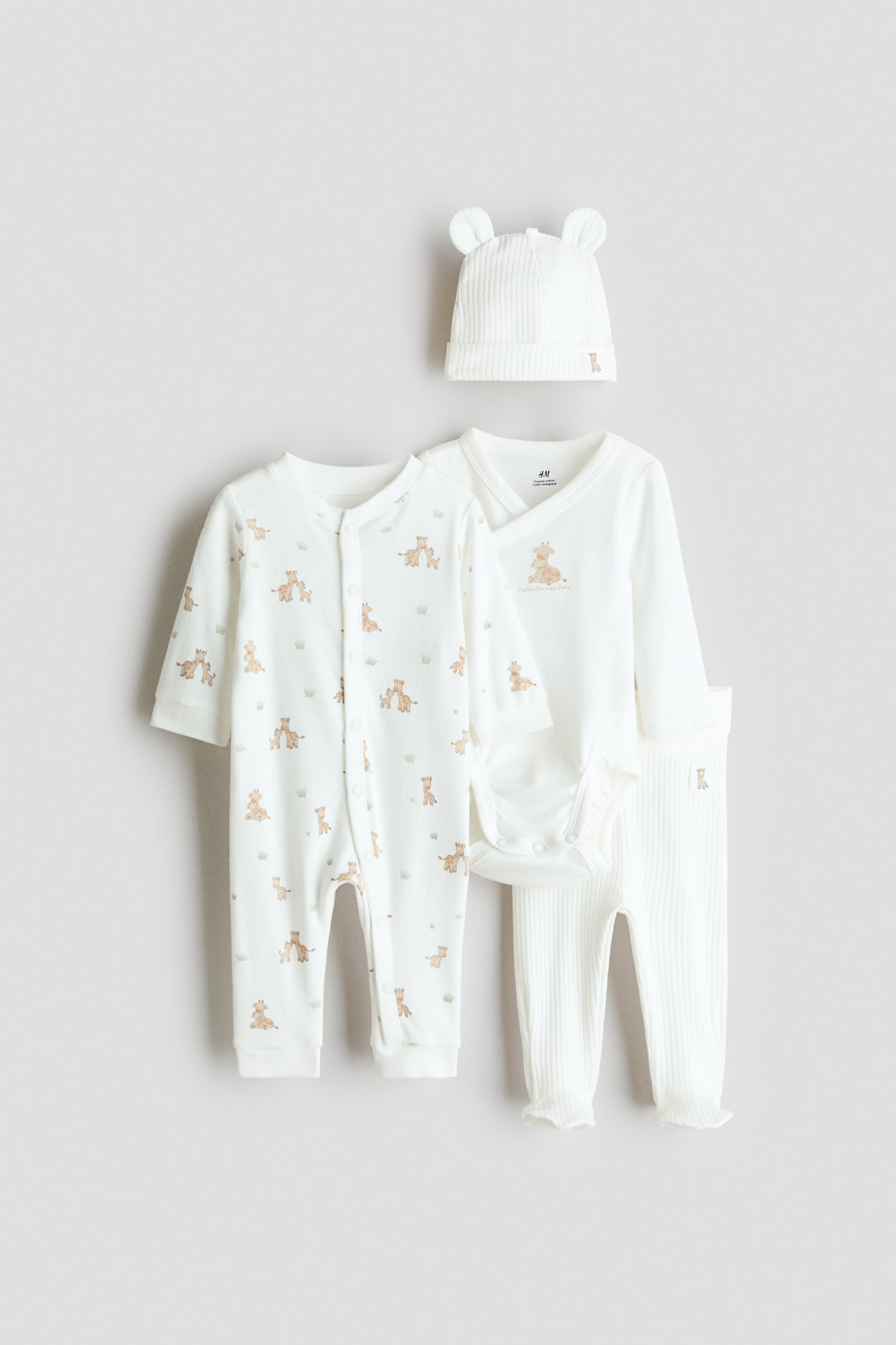 A four-piece cotton jersey baby gift set in off-white featuring a front-facing long-sleeve sleepsuit with an all-over giraffe print and snap-button fastenings. The set also includes a long-sleeve wrap bodysuit with a central giraffe graphic, ribbed knit leggings, and a coordinating ribbed beanie hat with small fabric ears.