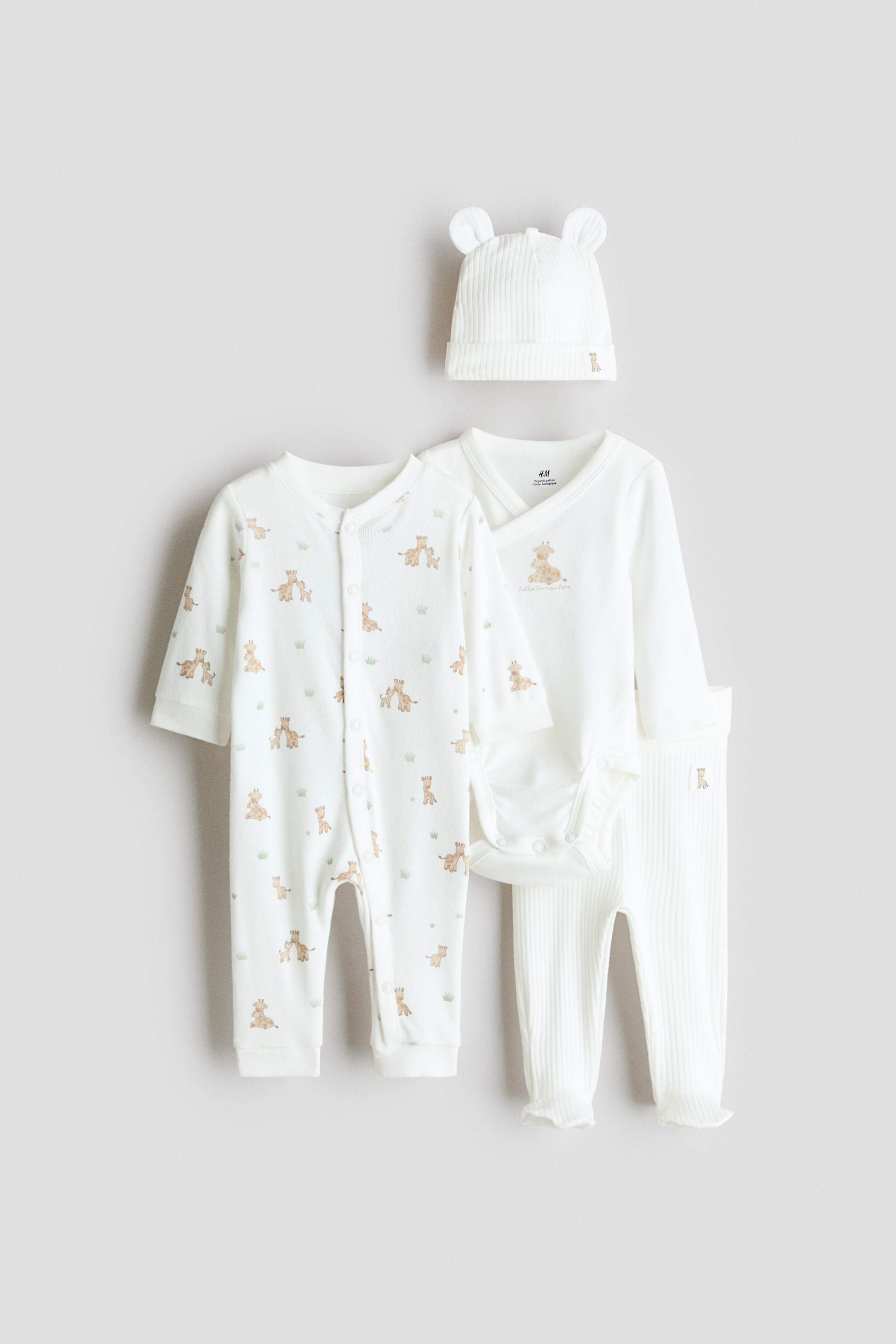 View larger image: A four-piece cotton jersey baby gift set in off-white featuring a front-facing long-sleeve sleepsuit with an all-over giraffe print and snap-button fastenings. The set also includes a long-sleeve wrap bodysuit with a central giraffe graphic, ribbed knit leggings, and a coordinating ribbed beanie hat with small fabric ears.