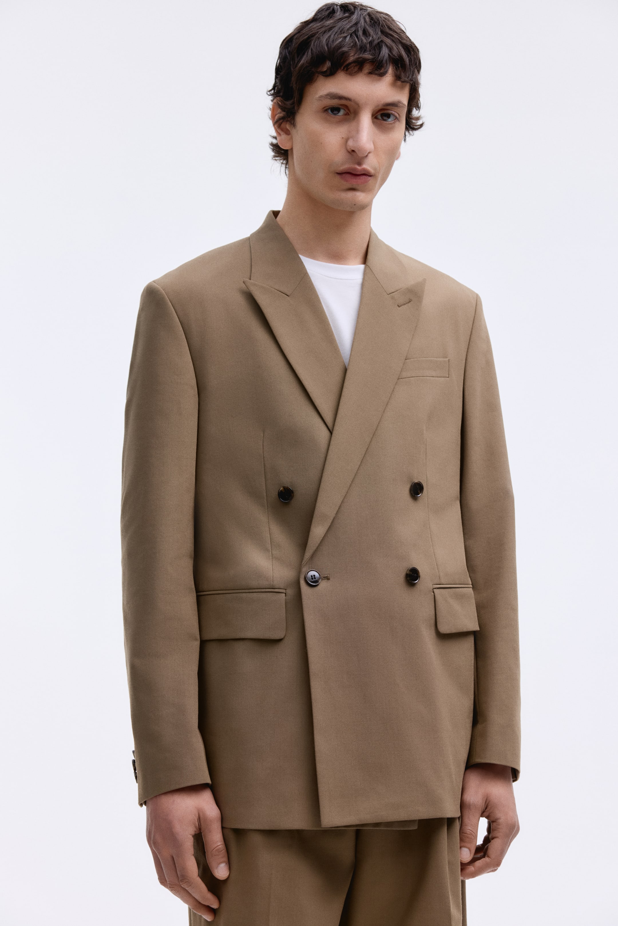Größeres Bild anzeigen: Ein hellbrauner, doppelreihiger Blazer wird von vorne gezeigt, mit breiten Notch-Revers, vier dunklen Knöpfen, zwei unteren Pattentaschen und einer oberen Leistentasche, darunter ist ein weißes Crew-Neck-T-Shirt sichtbar.
