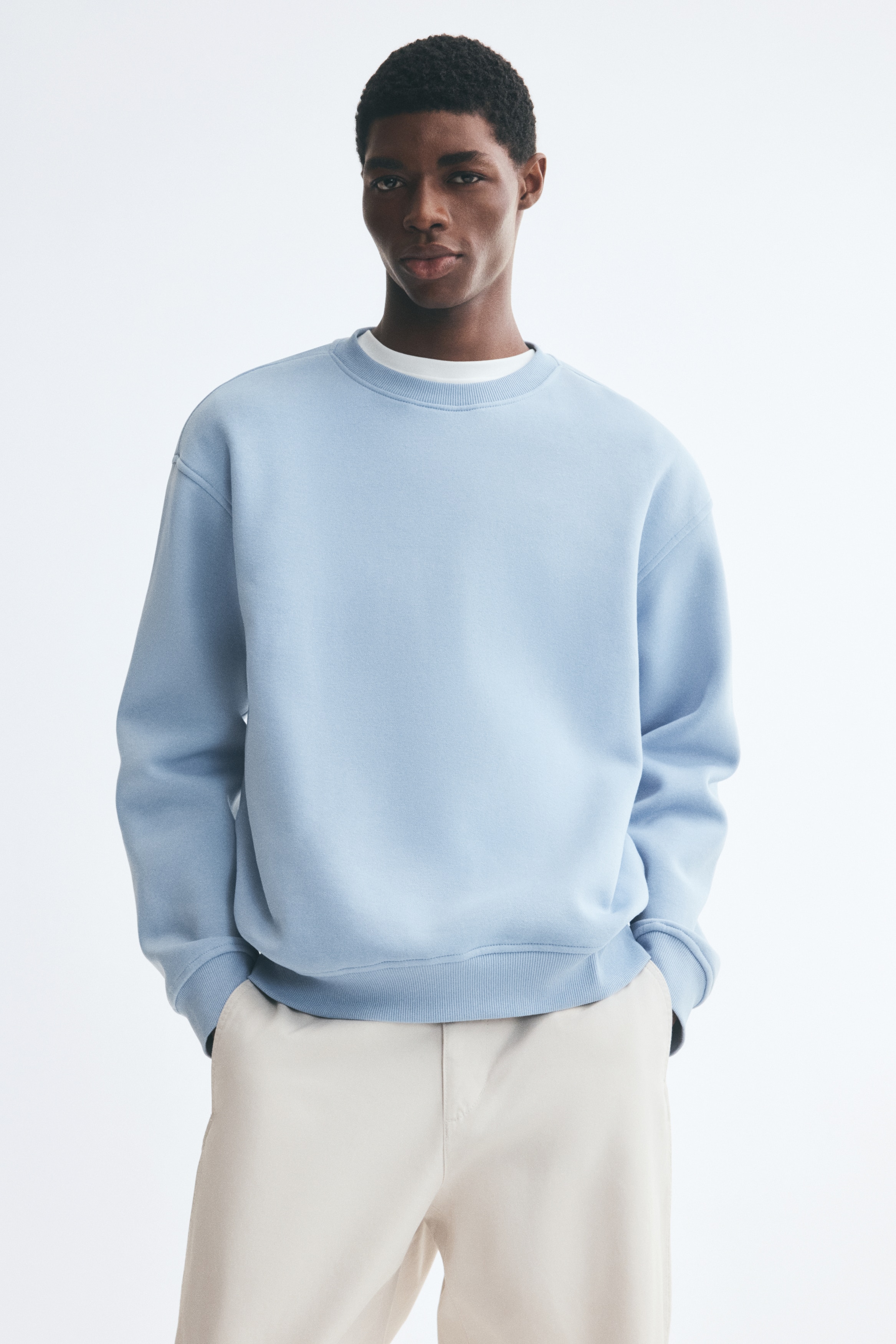 A light blue sweatshirt is hanging facing the front, showcasing a loose-fit silhouette with dropped shoulders and long sleeves. The garment features a round, rib-trimmed neckline alongside wide ribbed cuffs and a matching ribbed hem.