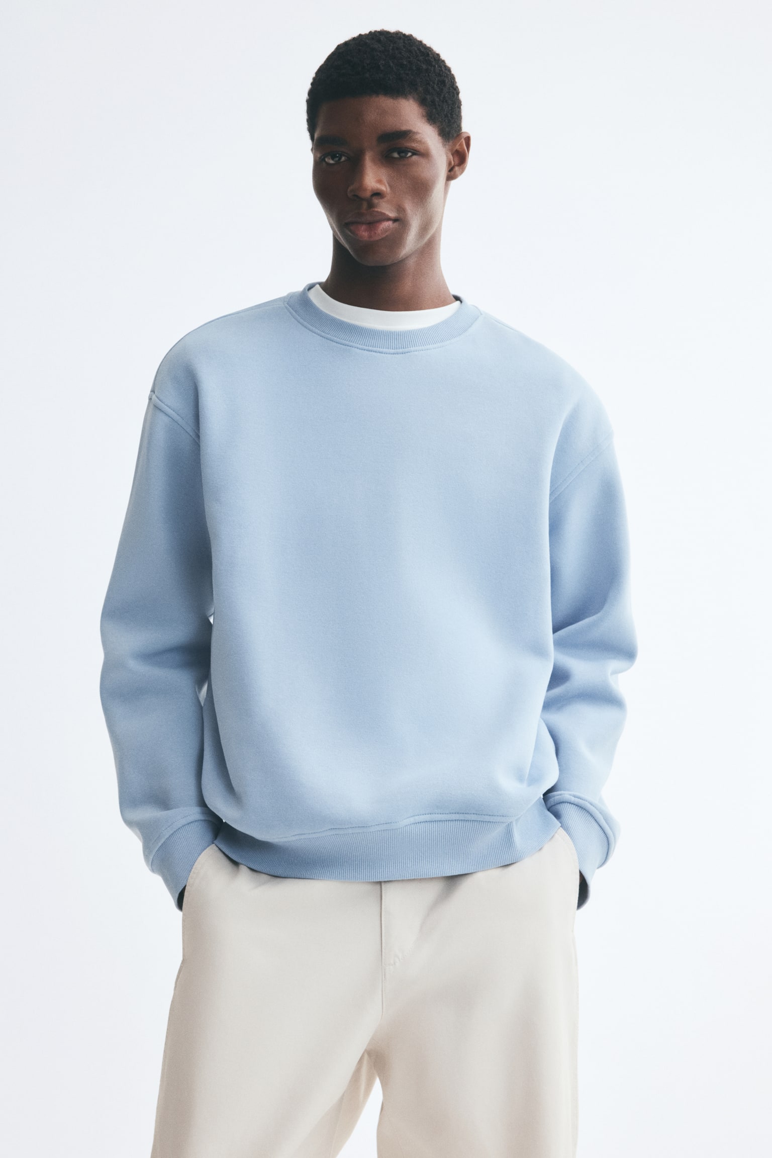 A light blue crewneck sweatshirt with a loose fit, dropped shoulders, and ribbed trims is being worn by the model over a white T-shirt. The model is standing with hands resting in the pockets of light beige trousers, causing the fabric of the sweatshirt to drape slightly at the hem. - 1
