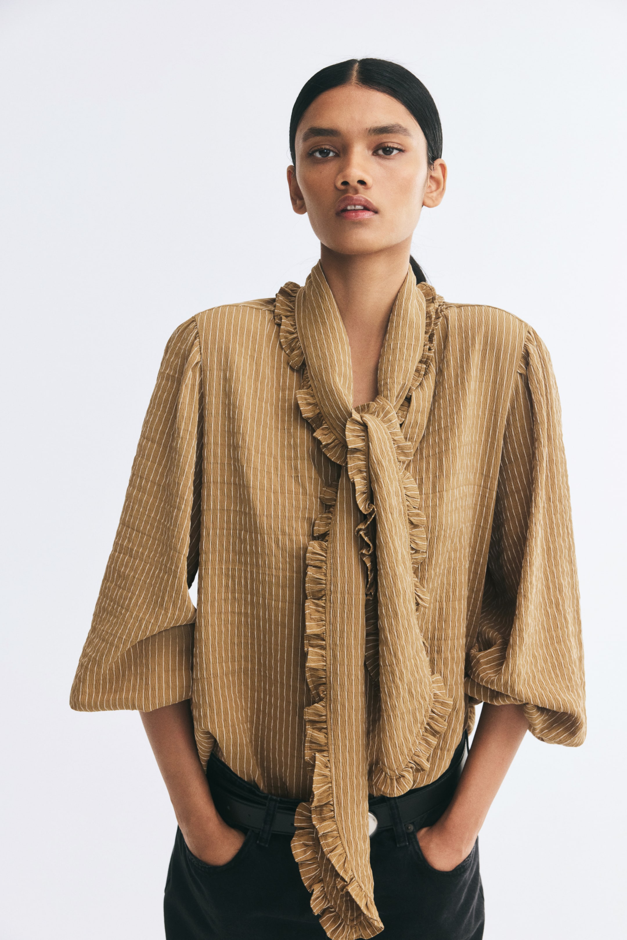 View larger image: A textured, ochre-coloured striped blouse is being worn by the model, featuring a ruffled scarf collar tied at the neck and long balloon sleeves. The model is posing with her hands placed in the pockets of black trousers.