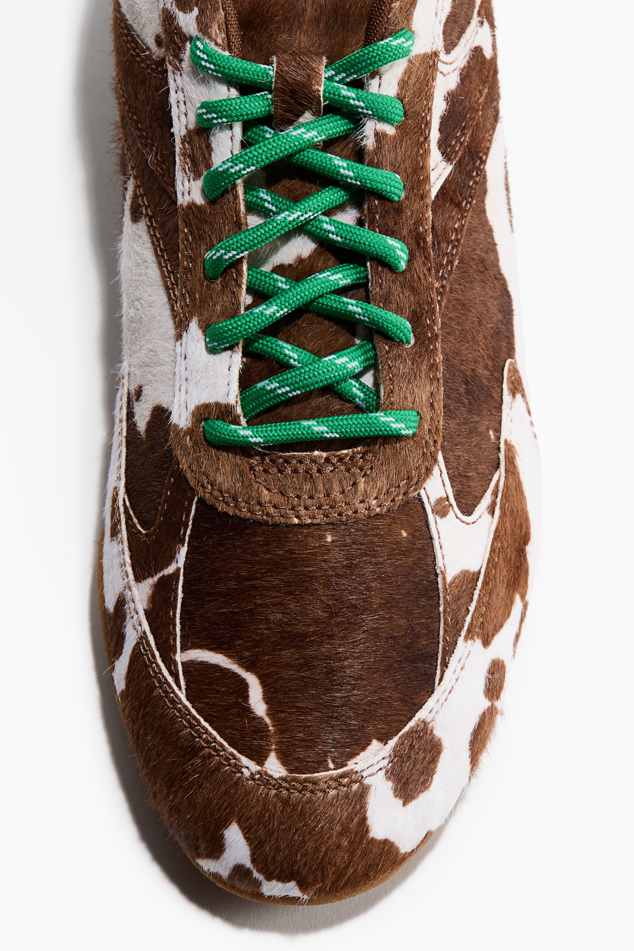 Ampliar la imagen: Un tenis muestra su parte frontal, con una textura peluda con estampado de vaca en marrón y blanco. Cordones redondos verdes con motas blancas se entrecruzan sobre la lengüeta.