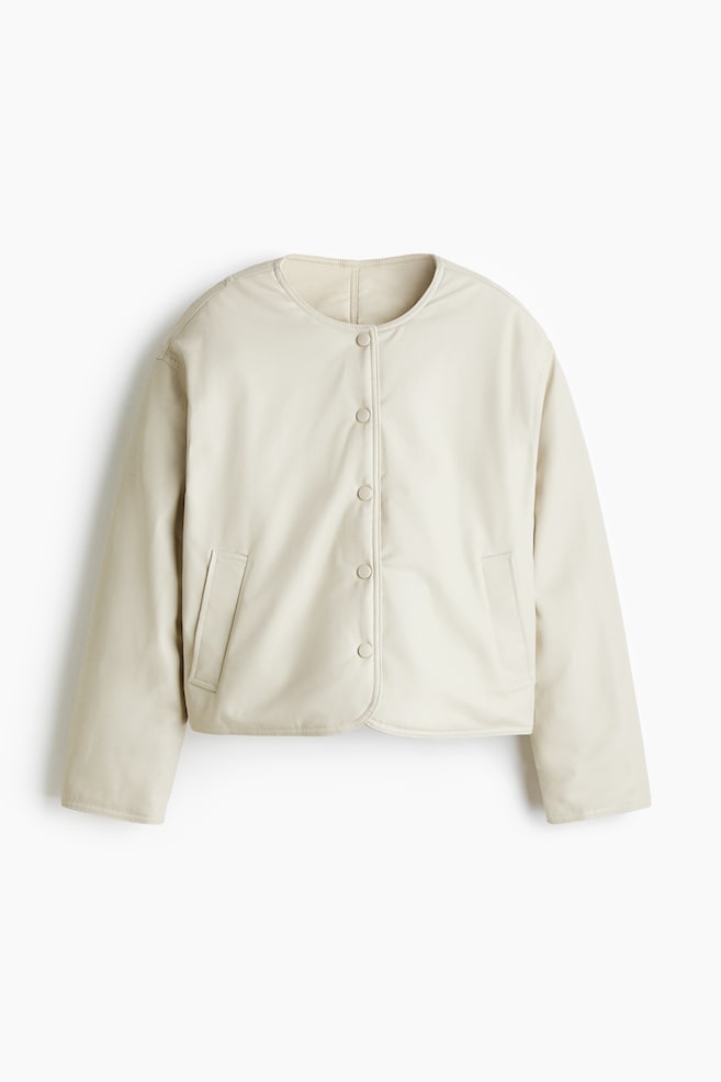 An off-white, collarless jacket is being presented from the front, showcasing a padded design with a smooth finish. It is featuring a full button-front closure with five visible buttons, two patch pockets, long sleeves, and a cropped, subtly rounded hemline. - 1