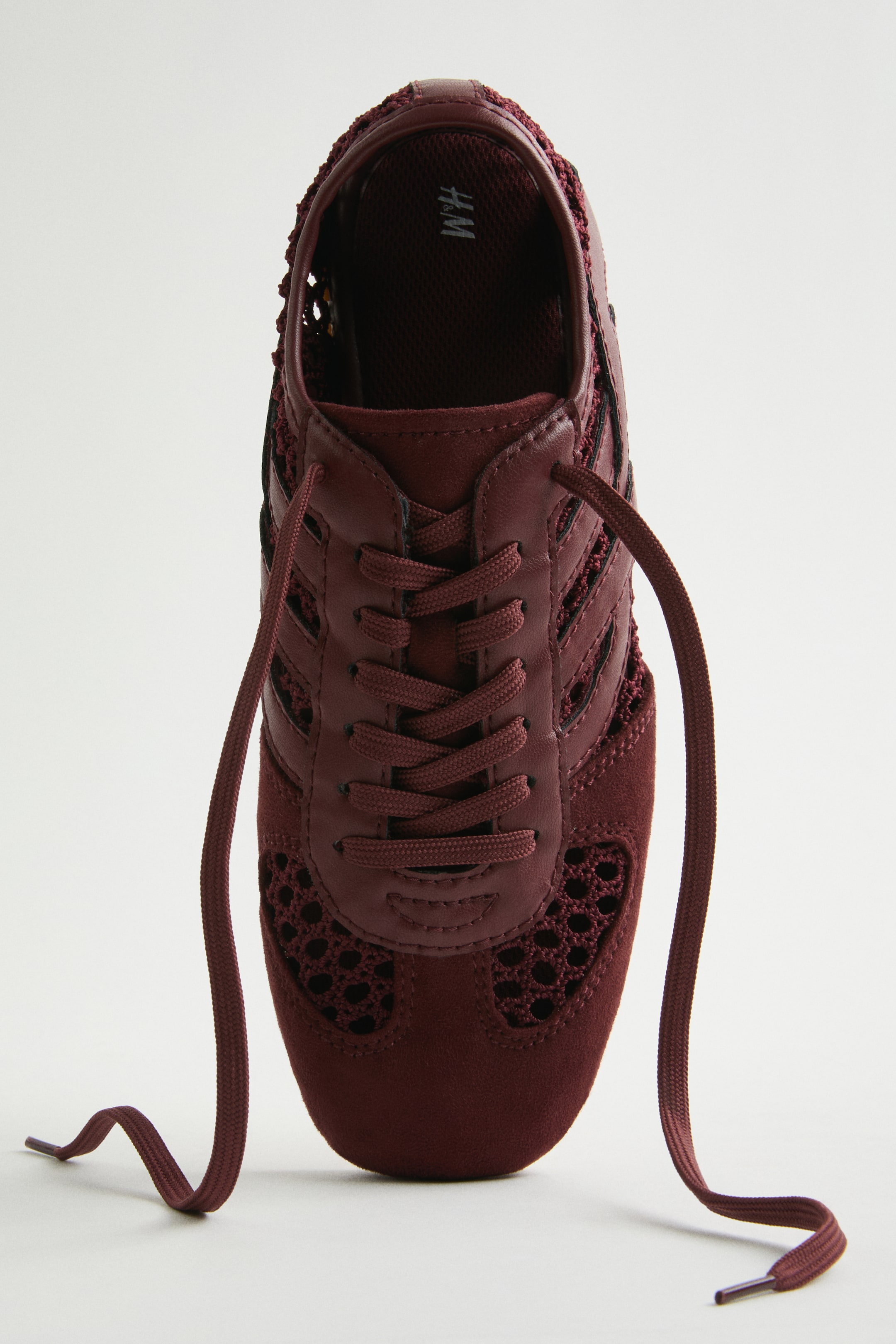 Grotere afbeelding bekijken: Een bordeauxrode sneaker staat rechtop en naar voren gericht, met de opening zichtbaar van bovenaf. Het toont gestructureerd suède en leerachtige panelen over geperforeerd gaas, en bijpassende losse veters hangen nonchalant langs de zijkanten.