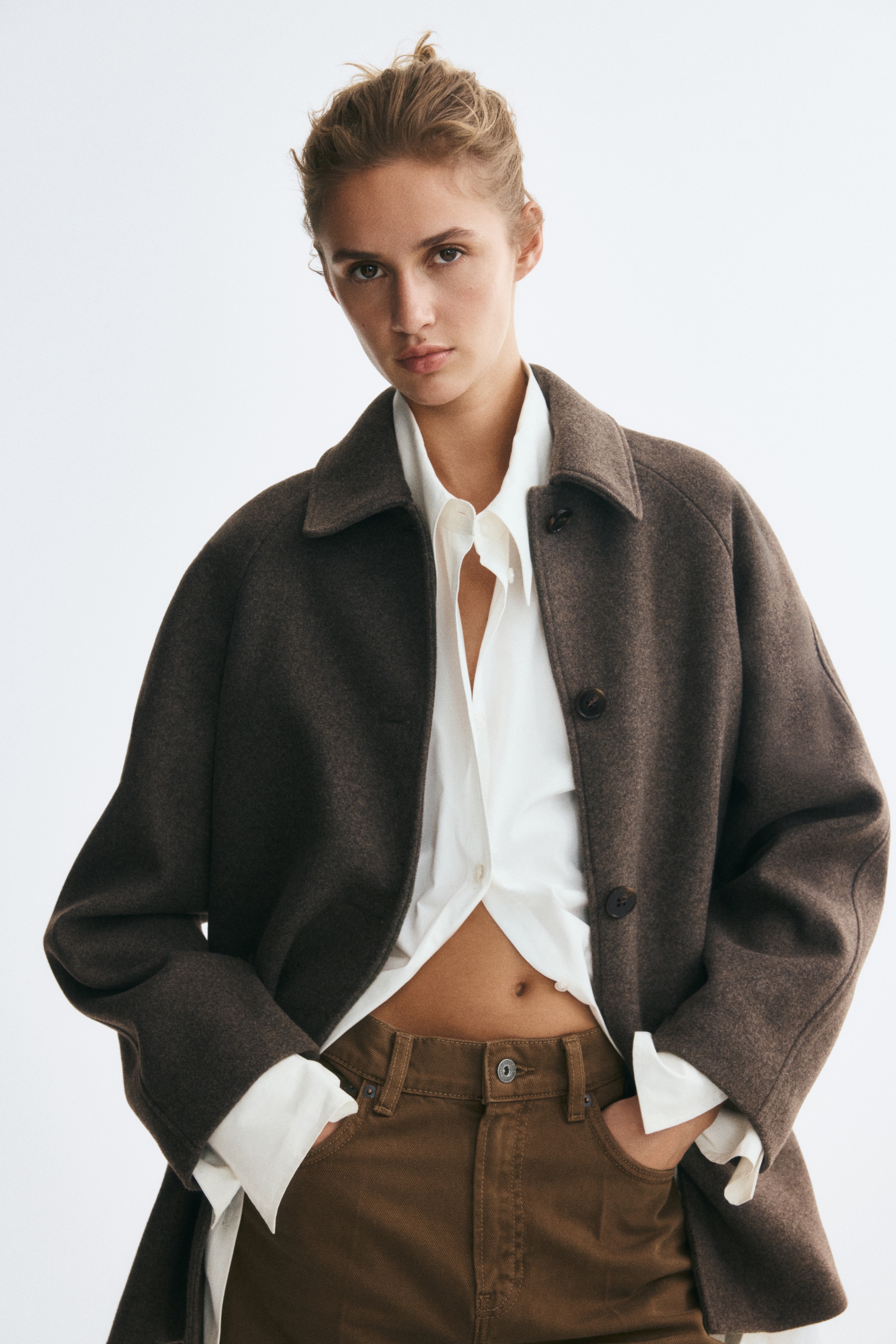 A dark brown, textured wool-blend coat is shown facing front, featuring a pointed collar, a button-front fastening with four dark buttons, raglan sleeves, and two front patch pockets with pointed flaps.
