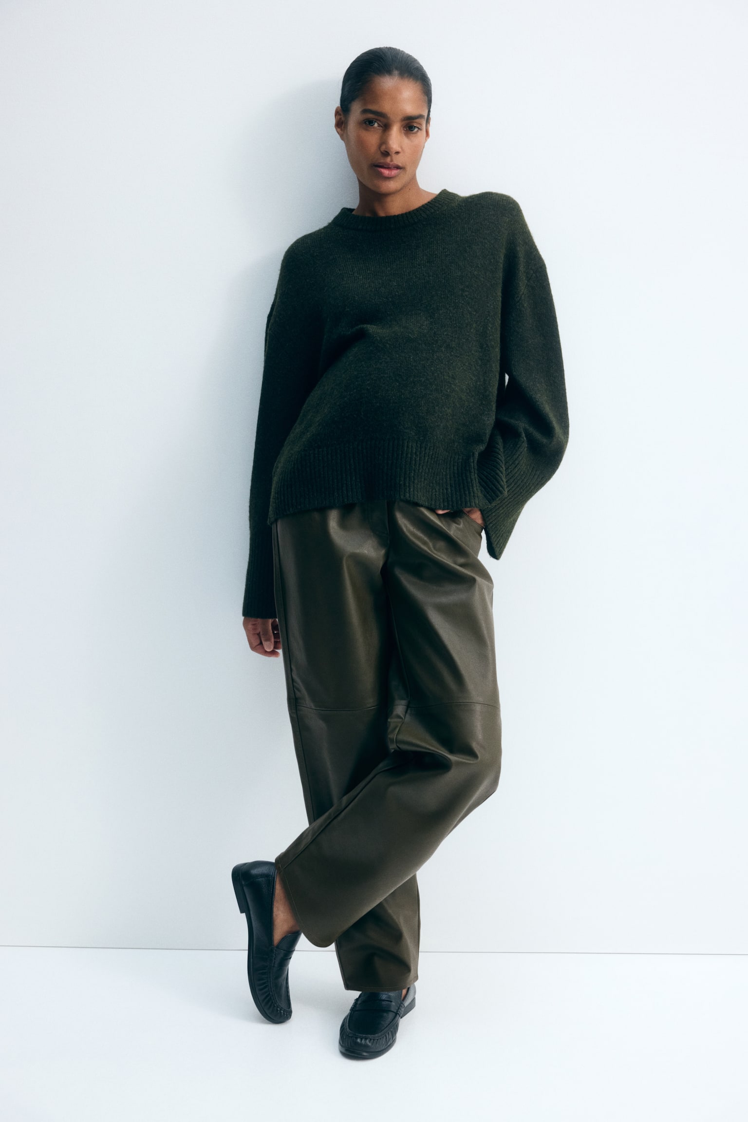 Bukser i coated stof i mÃ¸rk olivengrÃ¸n bÃ¦res af modellen og har en barrel-leg silhuet med vandrette sÃ¸mme ved knÃ¦ene. Modellen stÃ¥r med det ene ben krydset og den ene hÃ¥nd i en lomme og styler bukserne med en mÃ¸rkegrÃ¸n strikket sweater og sorte loafers. - 1