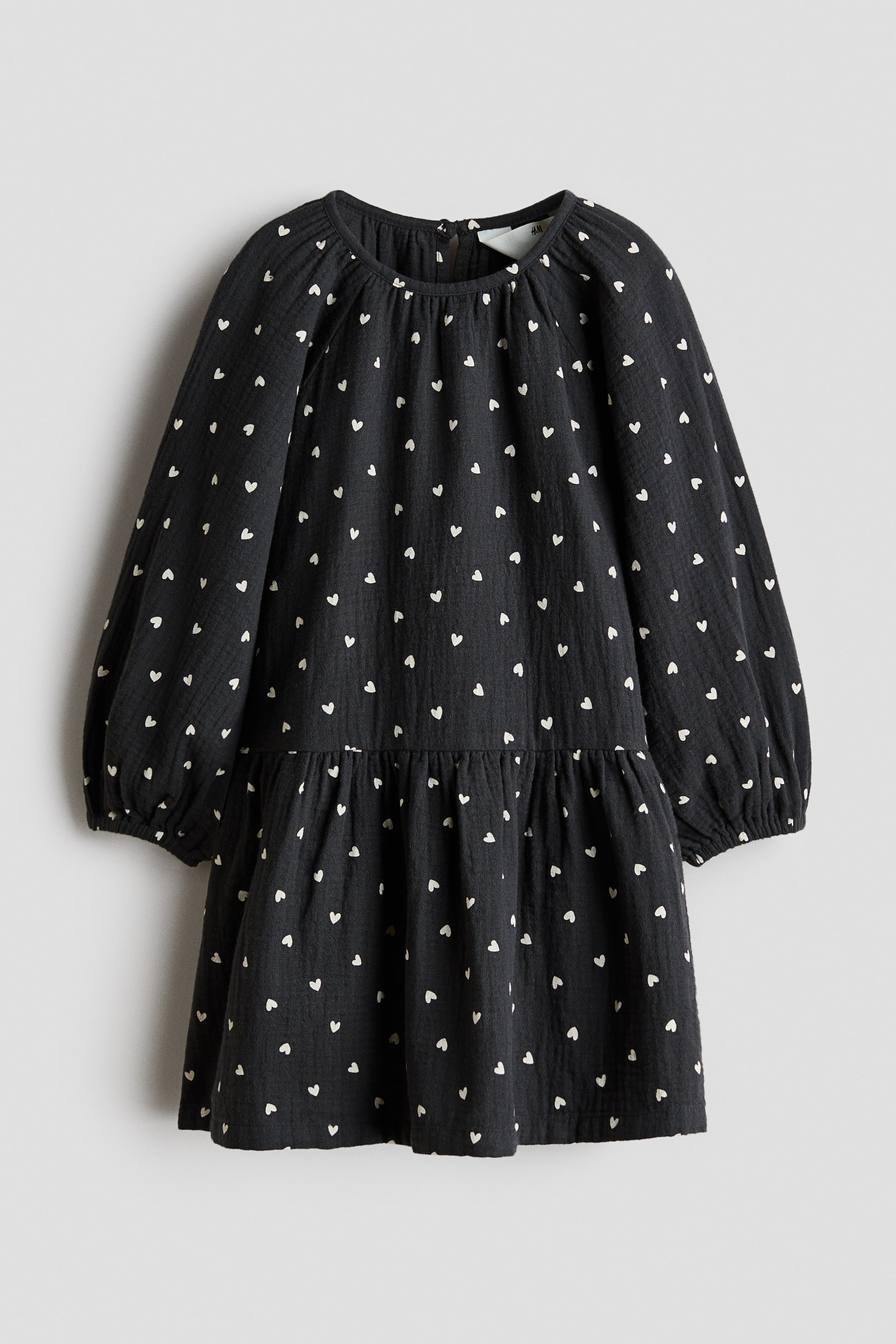 View larger image: A dark grey dress showcasing an all-over pattern of small white hearts. It is crafted with a round, gathered neckline, long voluminous sleeves with elasticated cuffs, and a gathered seam creating a tiered skirt.