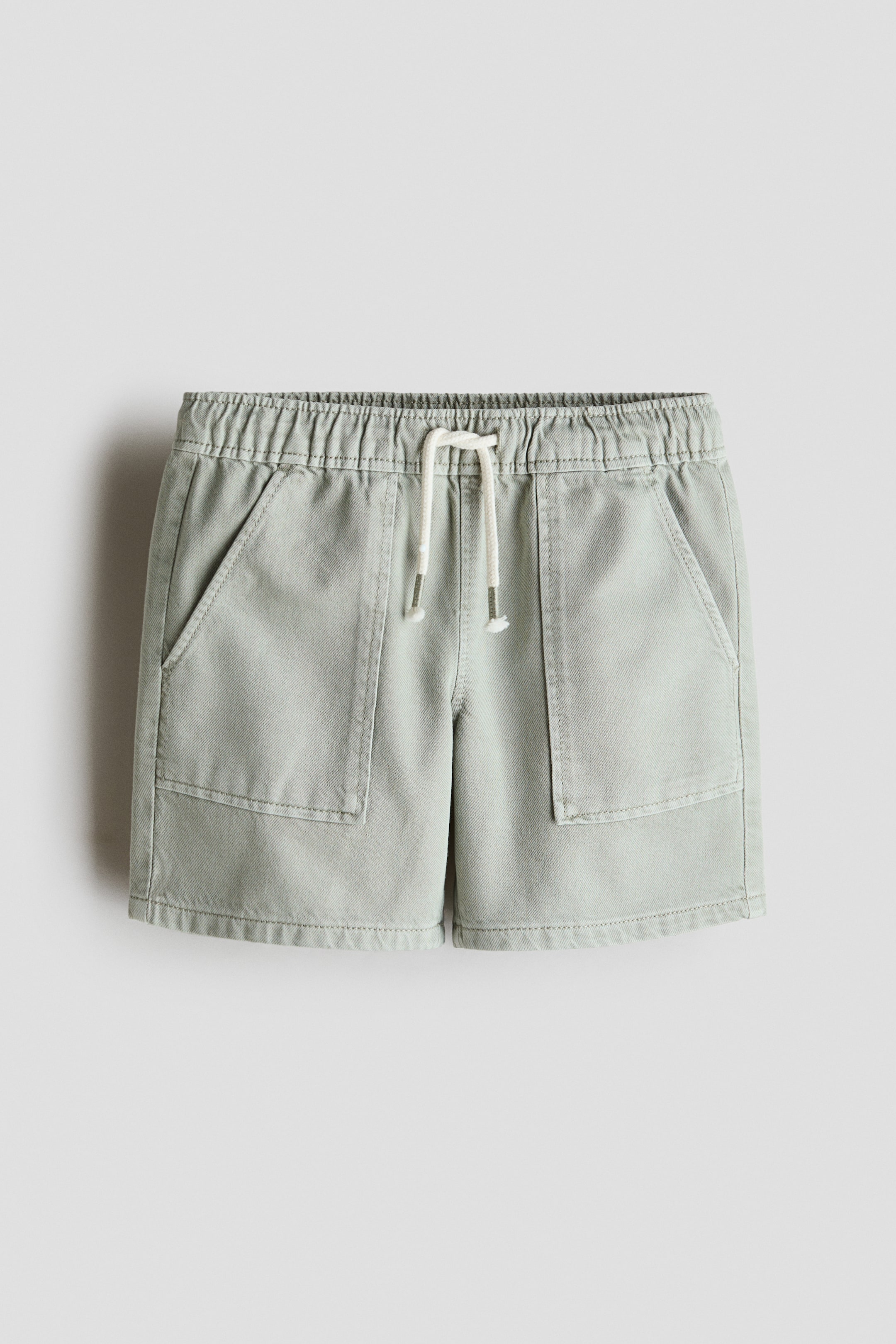 View larger image: Light green cotton twill shorts are showcased from the front, featuring an elastic drawstring waist with a white drawstring and two large front patch pockets.