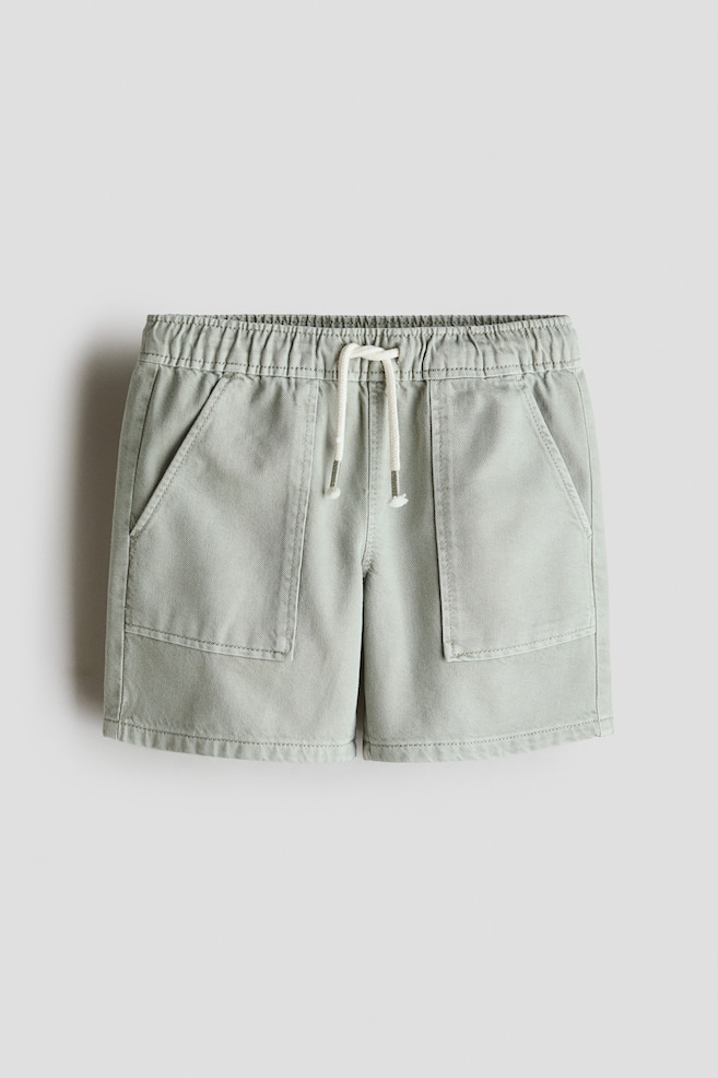 Shorts de sarga de algodón verde claro se muestran desde el frente, con cintura elástica ajustable con cordón blanco y dos grandes bolsillos frontales de parche. - 1