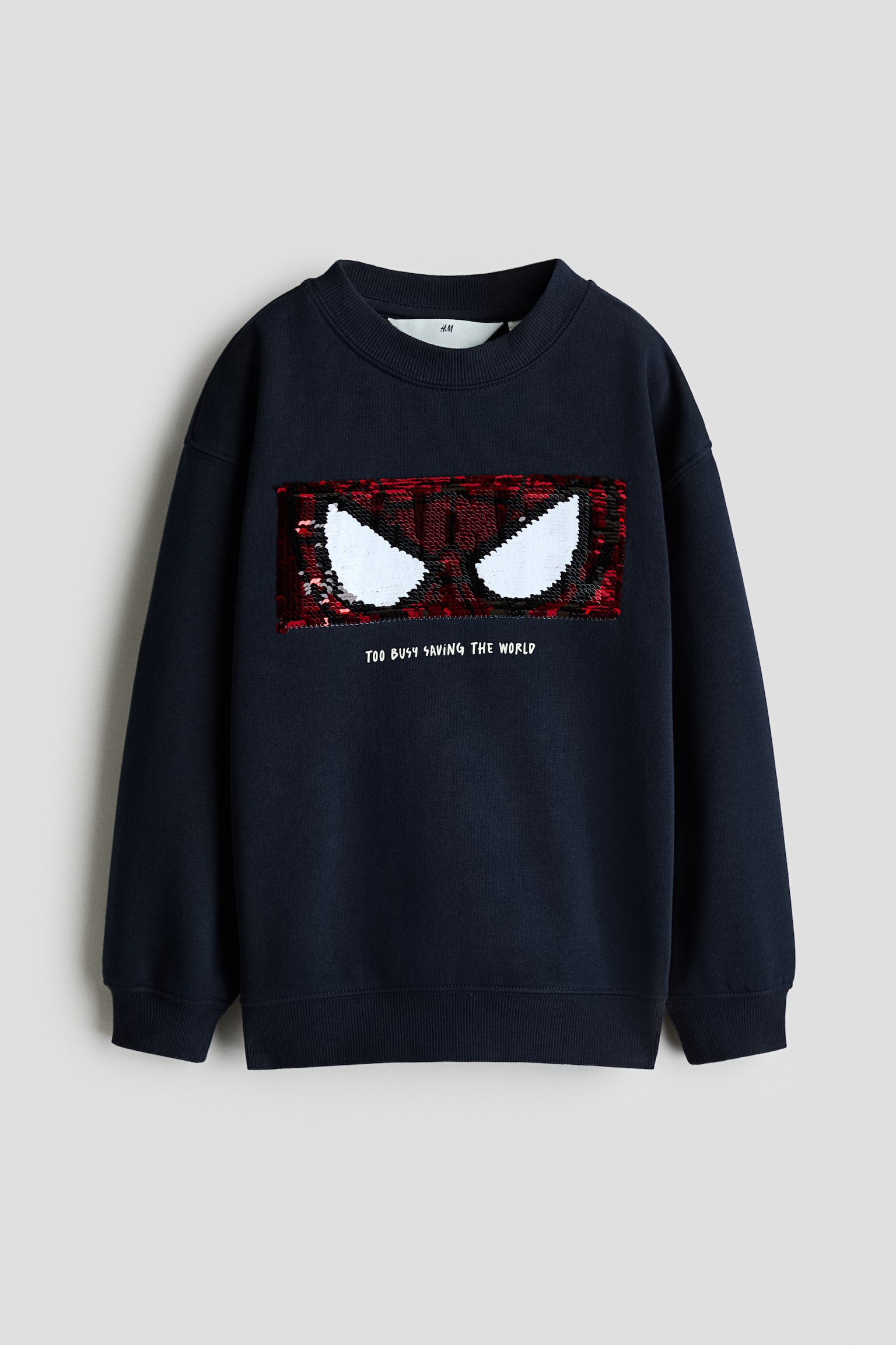 Un chandail Ã  col rond bleu marine foncÃ©, vu de face, affichant un motif rectangulaire en paillettes reprÃ©sentant les yeux de Spider-Man en rouge et blanc. Le chandail prÃ©sente des poignets et un ourlet cÃ´telÃ©s, avec le texte imprimÃ© blanc Â« TOO BUSY SAVING THE WORLD Â» placÃ© directement sous le motif en paillettes.