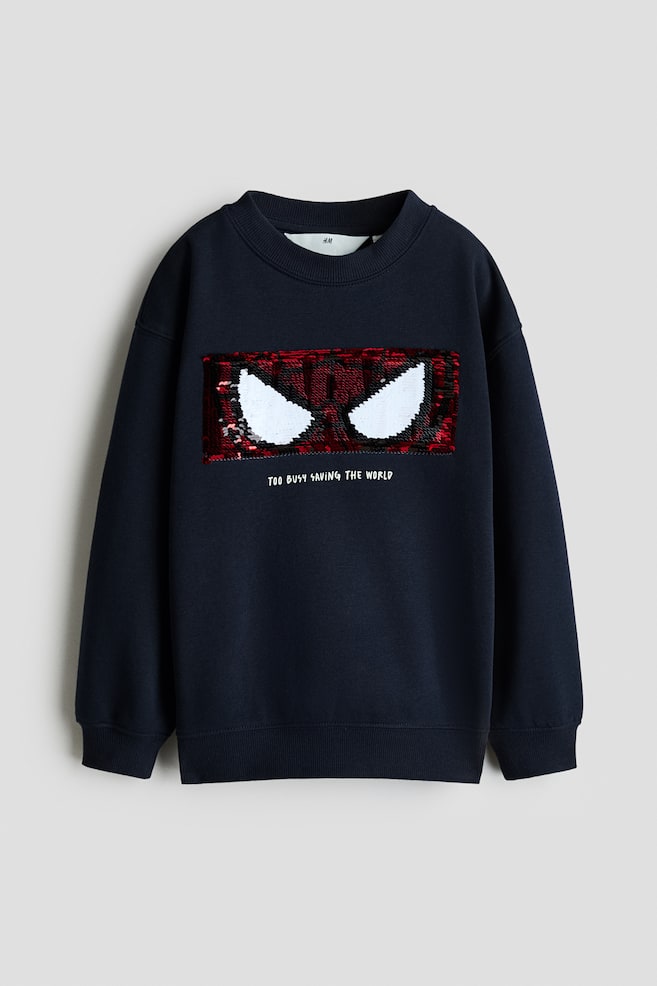 Una sudadera de cuello redondo azul marino oscuro, vista de frente, muestra un diseño rectangular de lentejuelas con los ojos de Spider-Man en rojo y blanco. La sudadera presenta puños y bajo acanalados, con el texto impreso en blanco "TOO BUSY SAVING THE WORLD" situado justo debajo del gráfico de lentejuelas. - 1