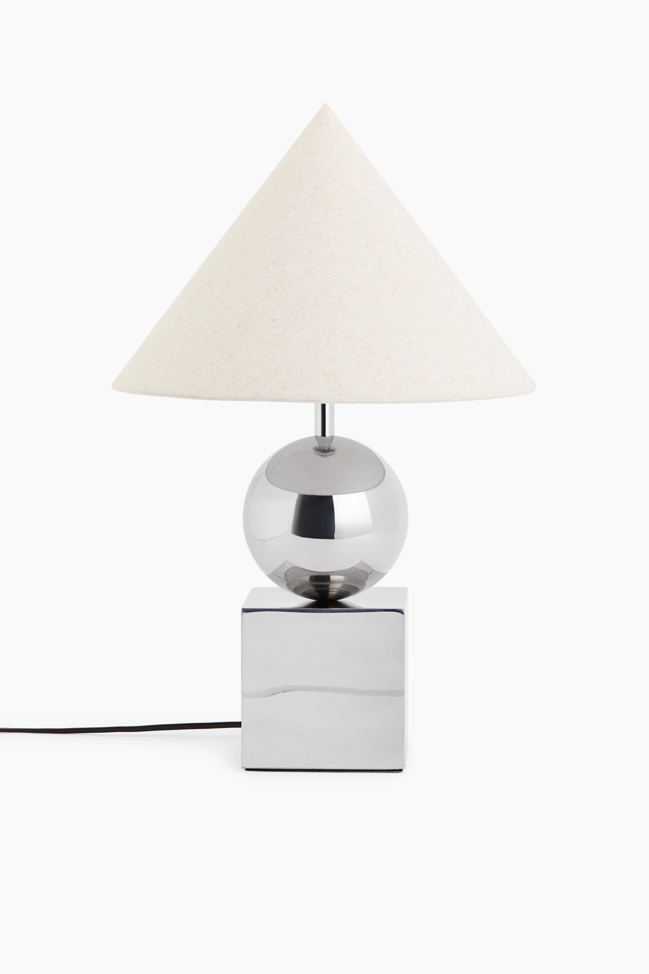 View larger image: The table lamp is showcasing its front view, featuring a conical, off-white fabric shade. This shade is resting on a slender chrome stem that emerges from a polished chrome sphere, which is positioned on a square, polished chrome base with a black power cable extending from its rear.