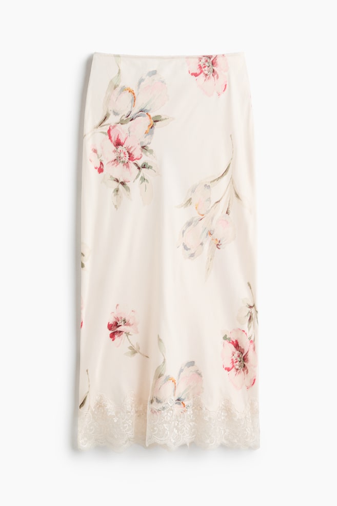 A cream-colored skirt is displaying a watercolor floral pattern with pink and red blossoms and green foliage. It is featuring a delicate, scalloped lace trim along its bottom hem. - 1