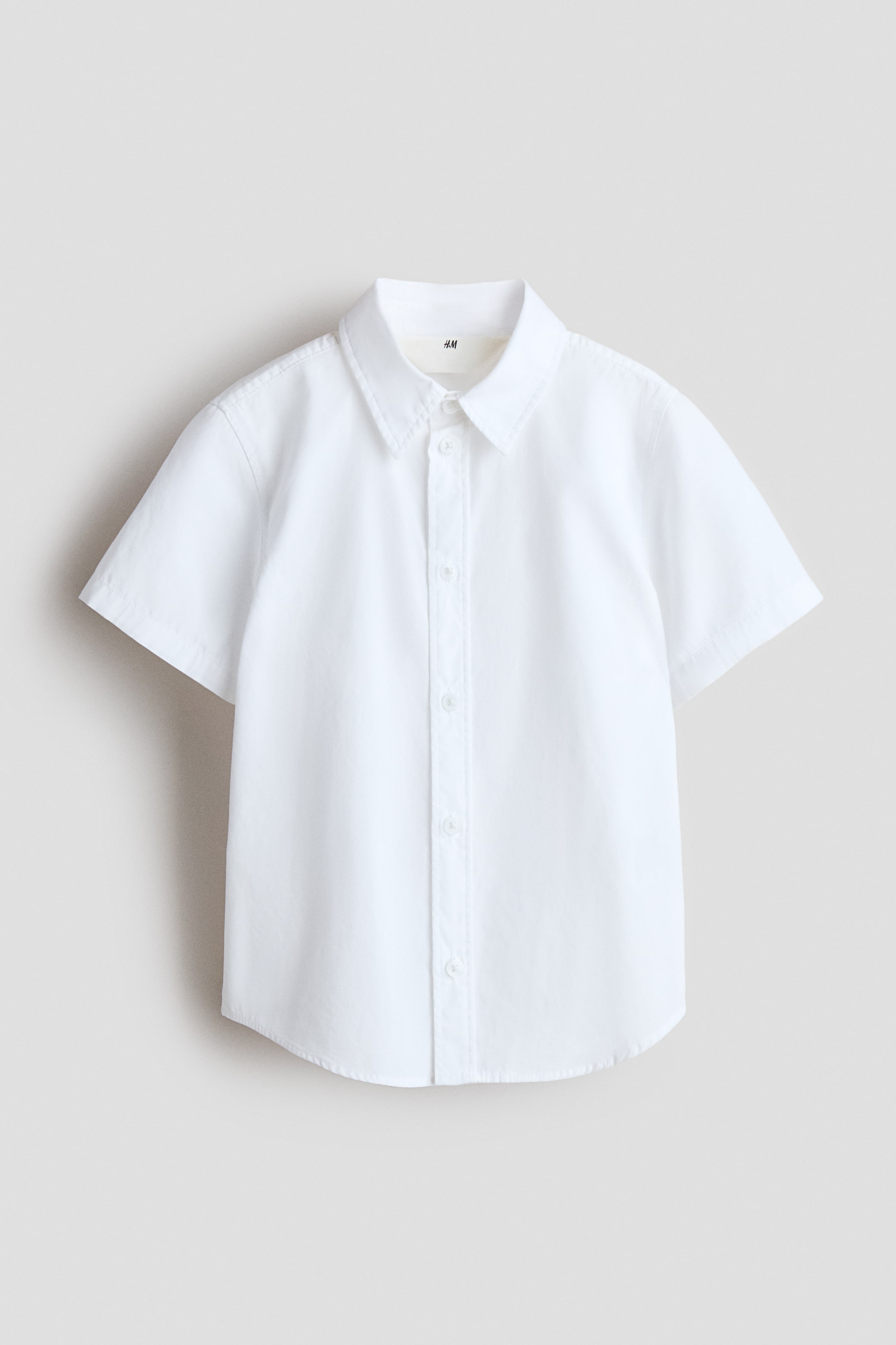 View larger image: A white short-sleeved shirt is presented FRONT, featuring a pointed collar, a full button-front closure with white buttons, and a rounded hem.