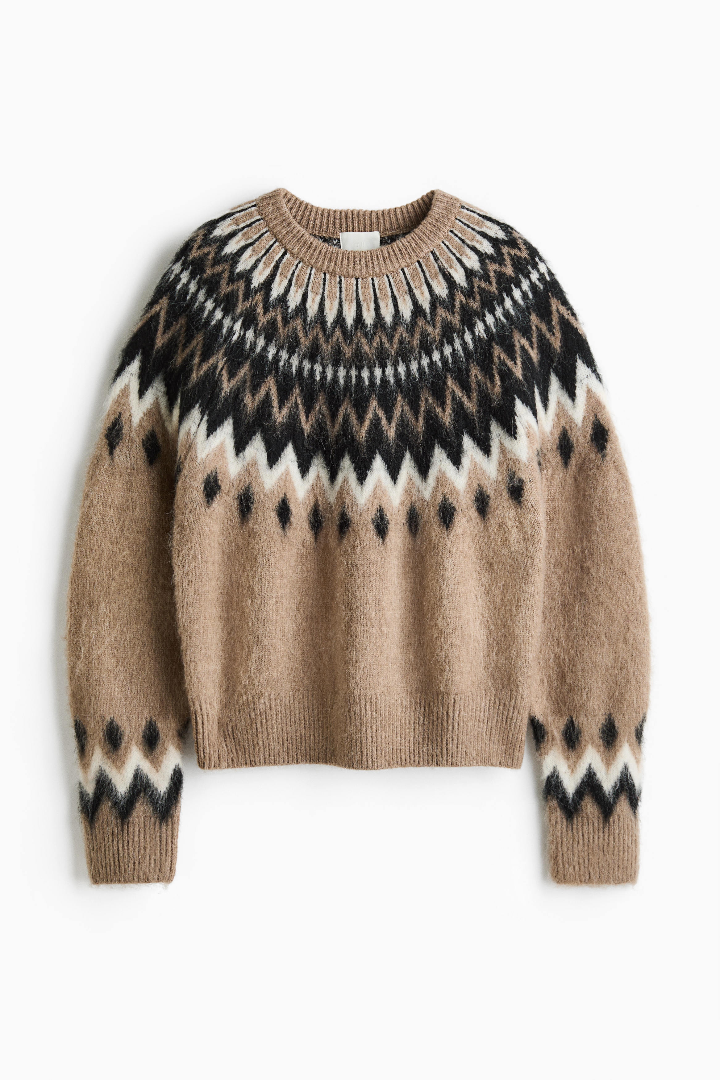 Un pull oversize en maille jacquard avec une finition brossée et un motif géométrique beige, noir et blanc est porté par le mannequin par-dessus un col roulé noir. Le pull présente un col rond bordé de côtes, de longues manches raglan ainsi que des poignets et un ourlet côtelés, et est associé à un pantalon noir.