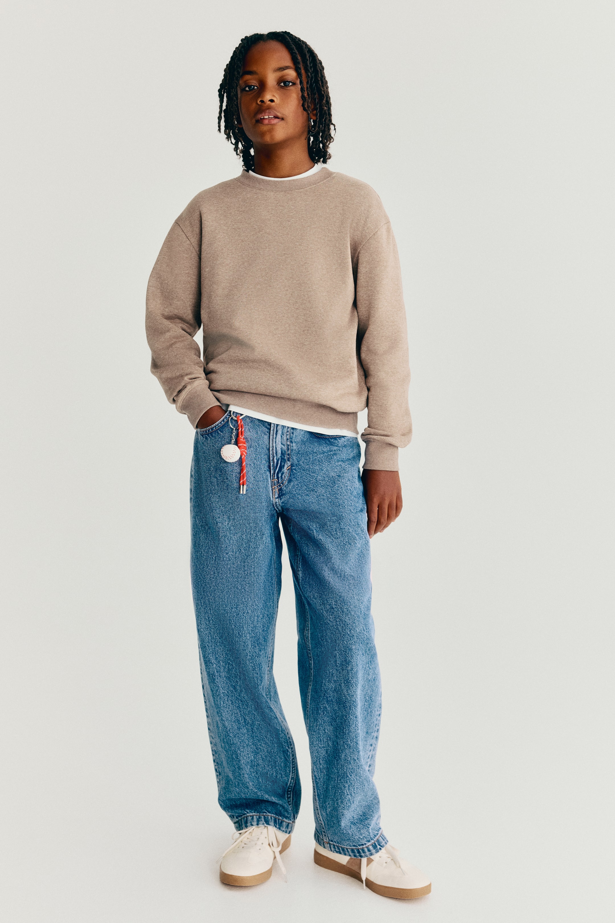 Se større billede: Disse 5-lommers jeans i vasket blå denim vises forfra og fremhæver en løs pasform over hofte og lår med lavt skridt og vide ben. Jeansene styles med en beige sweatshirt og hvide sneakers, med en lille baseballformet charm hængende fra den forreste bæltestrop.