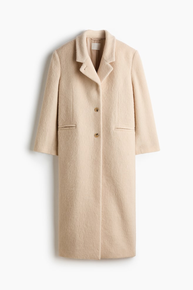 A long, single-breasted coat in a textured, light-beige fabric is hanging facing front, showcasing notch lapels and a two-button closure. The coat is displaying a straight silhouette with long sleeves and two horizontal welt pockets at the waist. - 1