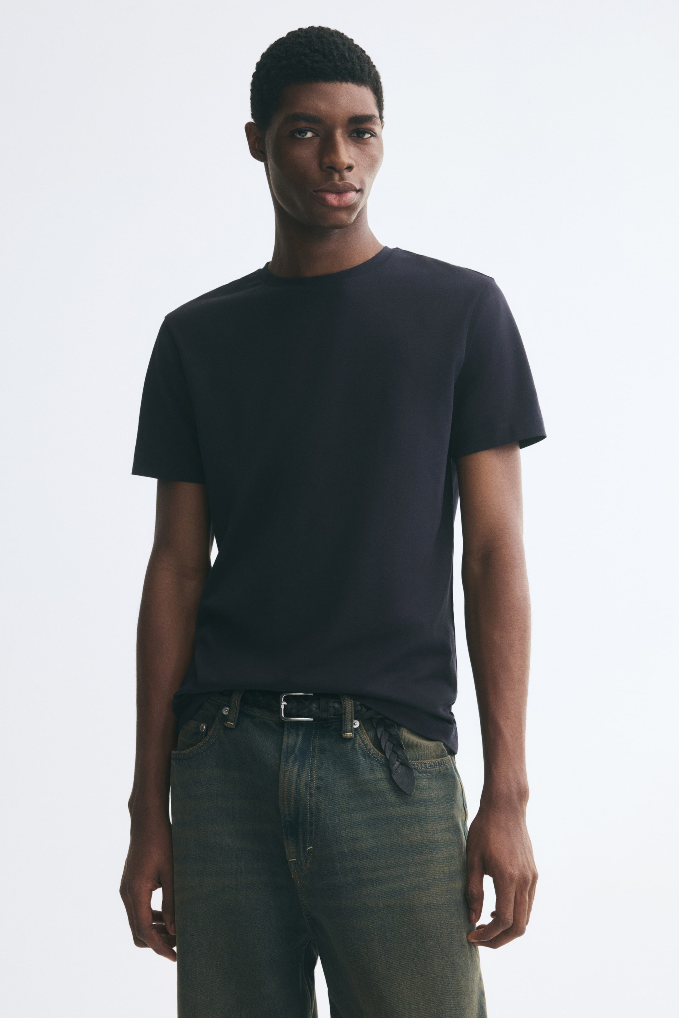Größeres Bild anzeigen: Das Model trägt ein dunkel marineblaues T-Shirt mit Rundhalsausschnitt und kurzen Ärmeln, das leicht über den Bund einer ausgewaschenen Jeans in Olivton fällt.