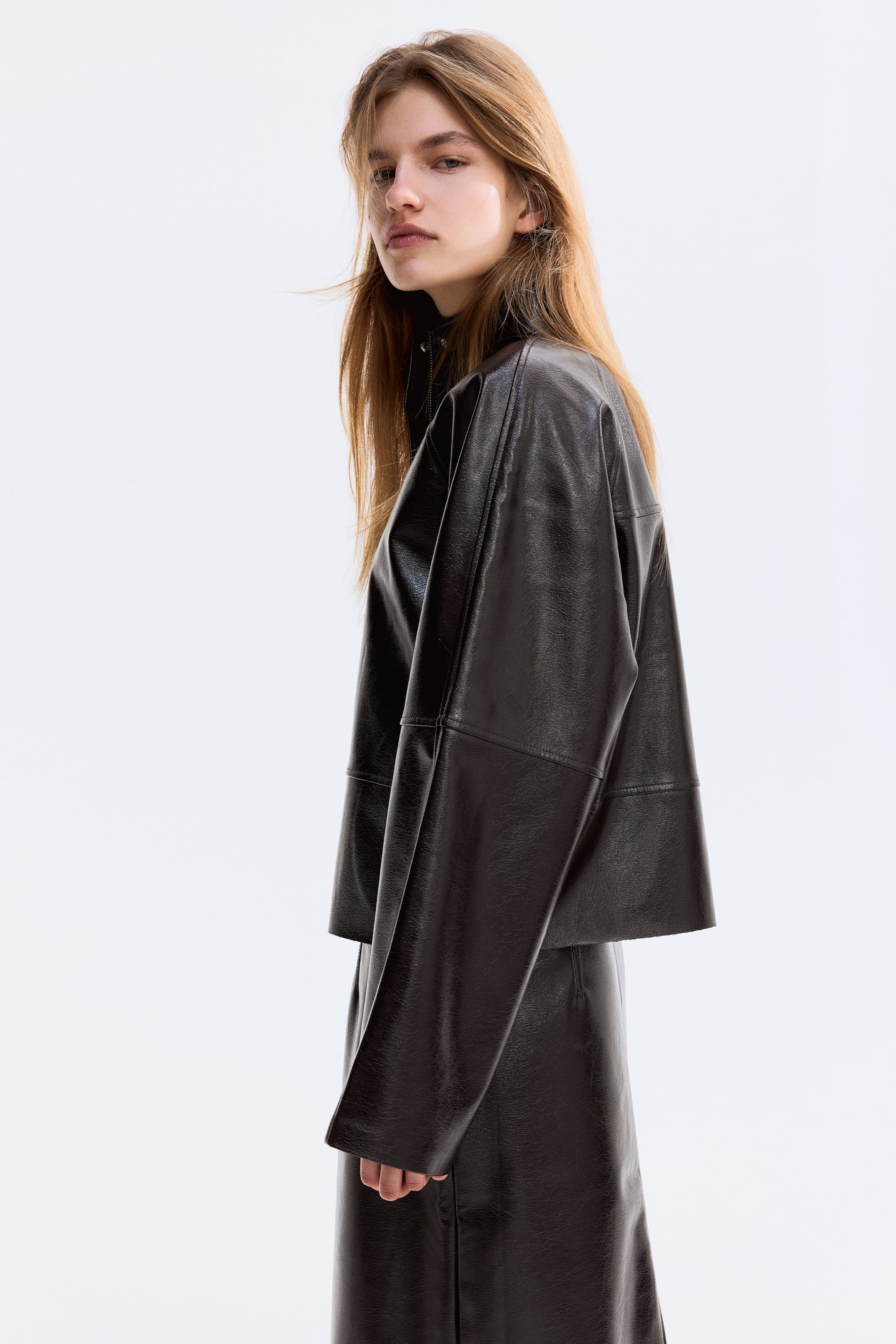 Agrandir l'image: Une veste sombre, texturée et brillante est présentée sous un angle de côté-avant, avec un col haut, des manches larges et droites, et un ourlet court. Une jupe sombre assortie est visible en dessous, complétant l'ensemble.