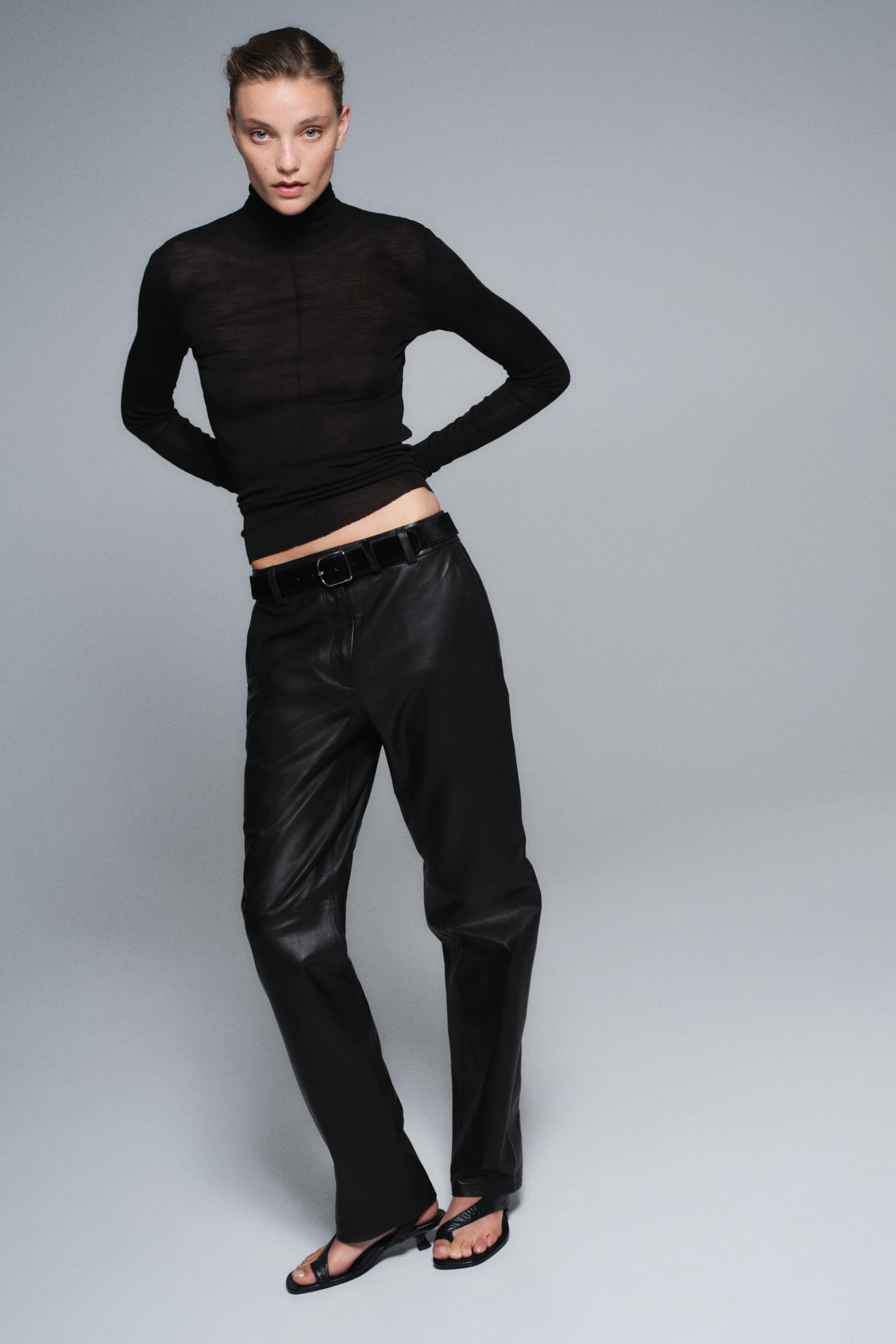 Ver imagen más grande: Estos pantalones negros de cuero se presentan de frente, con una silueta de pierna recta y una cintura de tiro medio ajustada con un cinturón negro a juego. Se combinan con un top negro de cuello alto, de manga larga y semitransparente, que realza su corte relajado pero estructurado.