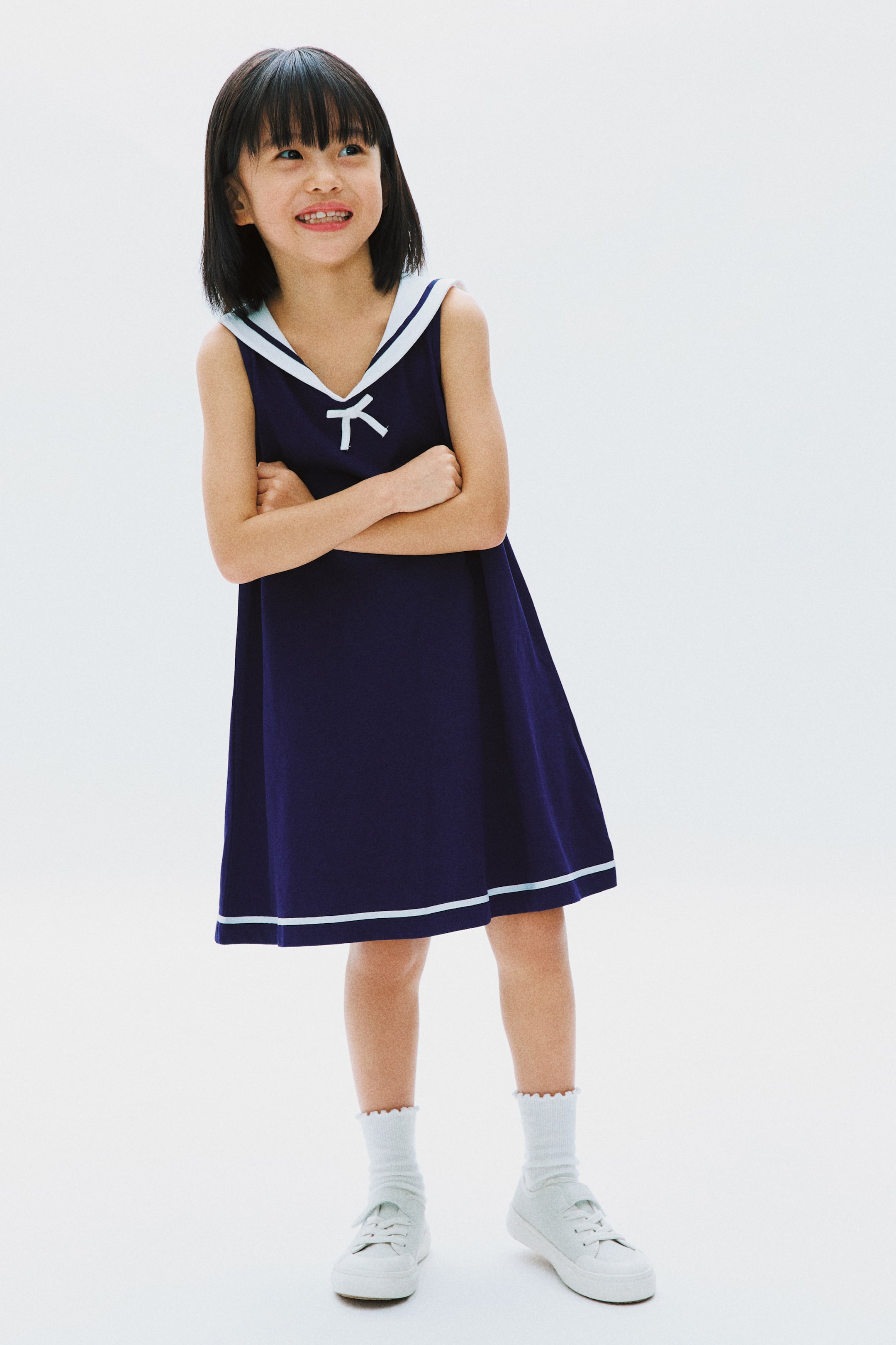 View larger image: A navy blue sleeveless dress featuring a white sailor collar with a V-neck and a small white bow, complemented by two thin white and light blue stripes running above the hemline.