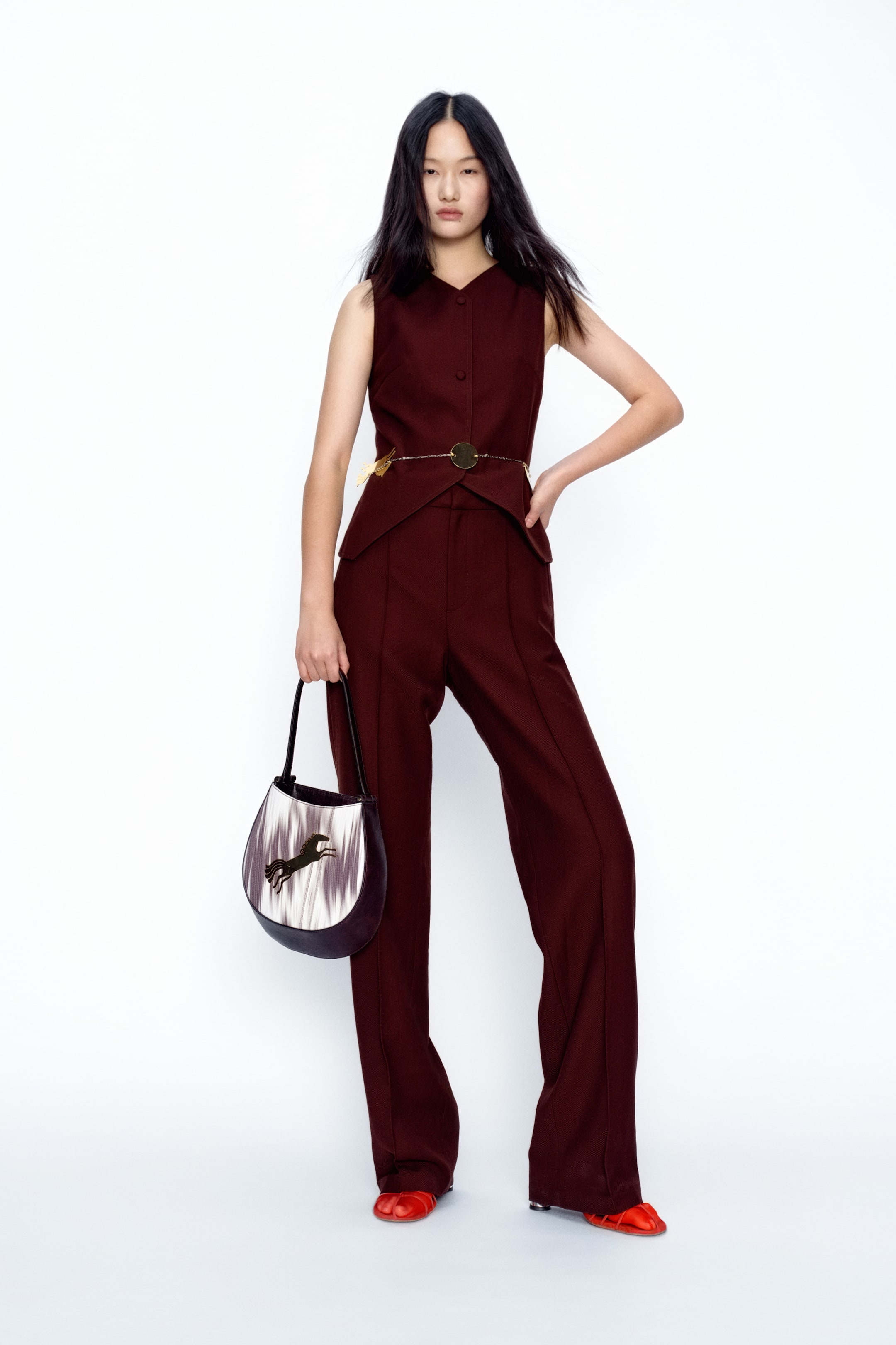 View larger image: A dark red sleeveless V-neck top with button-front closure and a peplum hem is paired with matching wide-leg trousers featuring a front seam. A gold-tone chain belt with a circular buckle cinches the waist, while a structured dark red and white bag with a horse graphic and red split-toe flats complete the look.
